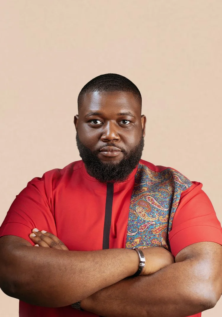 Portrait of a man with a beard, crossing his arms, wearing a red shirt with a paisley design on one shoulder, against a plain beige background.