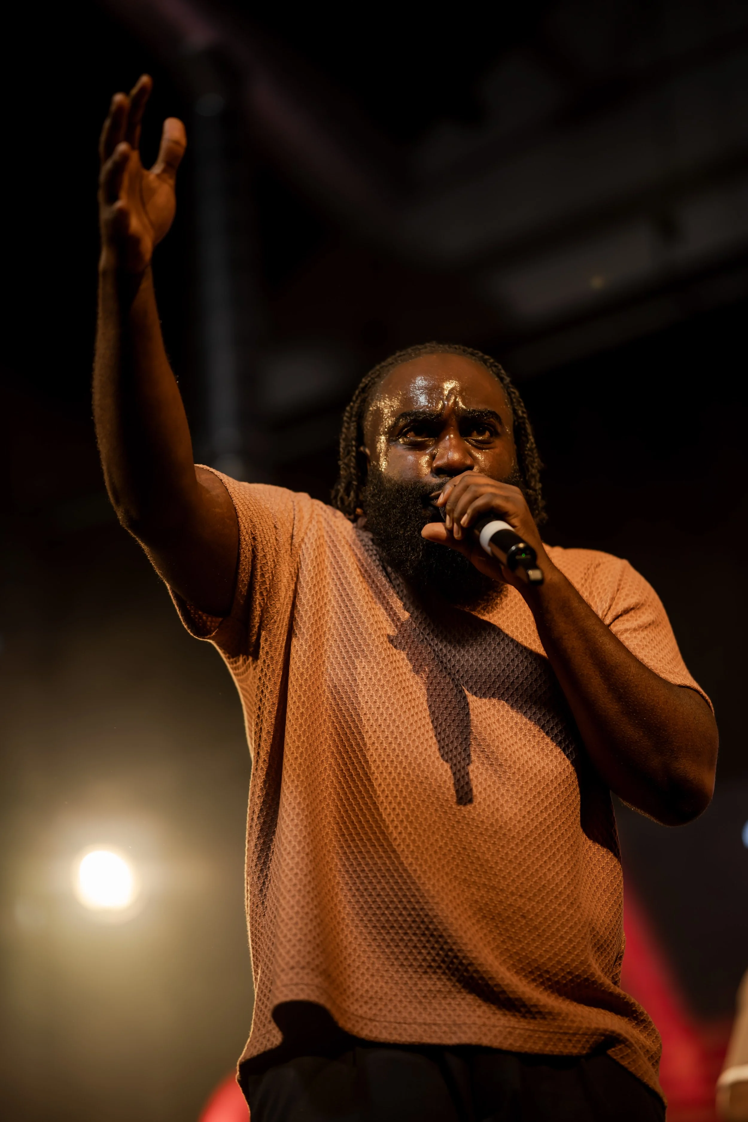 A man with dreadlocks and a beard is singing into a microphone on stage. He is wearing a peach-colored textured shirt and raising his right hand. The stage is dimly lit with a bright light behind him.
