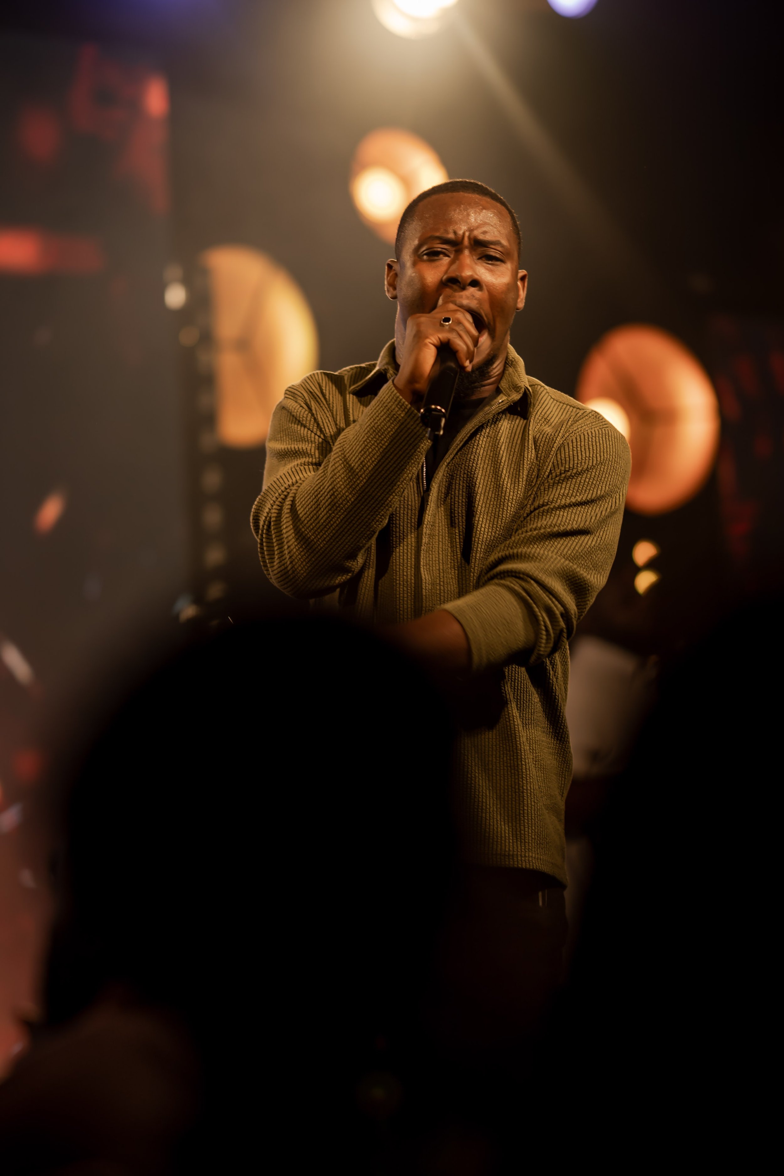A man singing into a microphone on stage with warm lighting and blurred circular lights in the background.