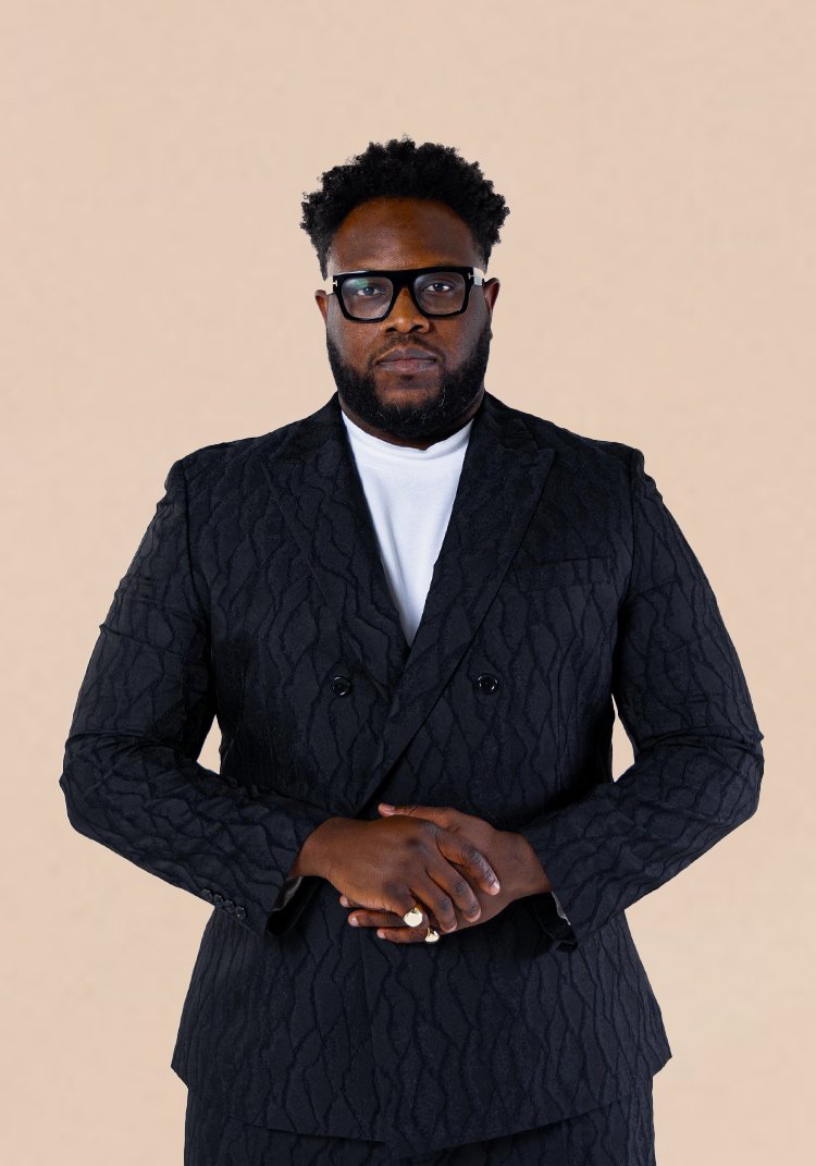 A man with dark skin, black curly hair, and a full beard wearing black glasses, a white t-shirt, and a black textured double-breasted blazer standing against a plain light beige background.