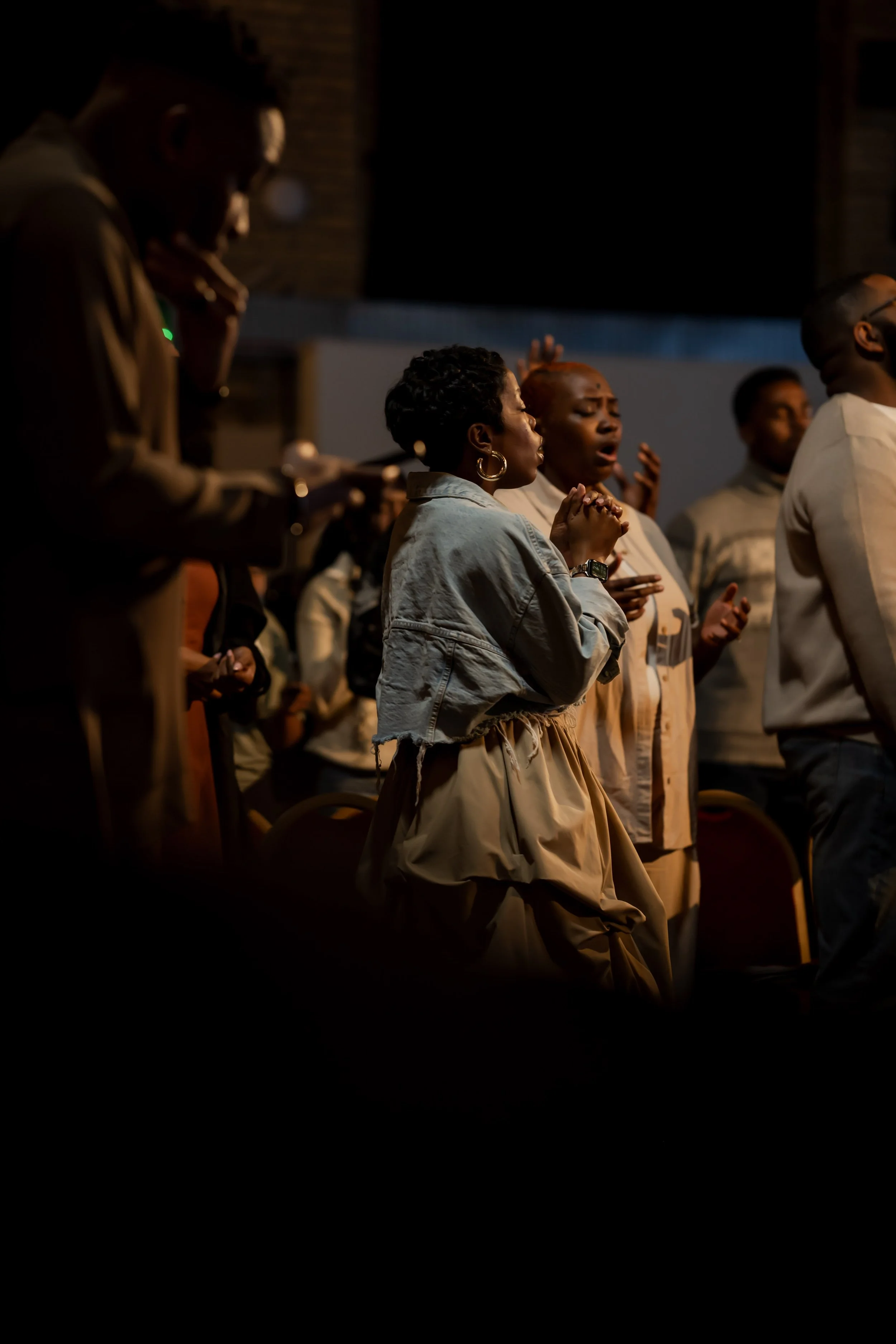 People praying or singing with eyes closed in a dark setting, some with hands raised.