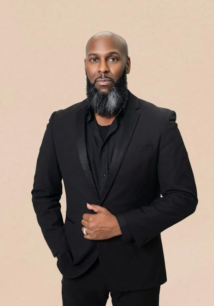 A bald man with a beard wearing a black suit and shirt, standing against a beige background.