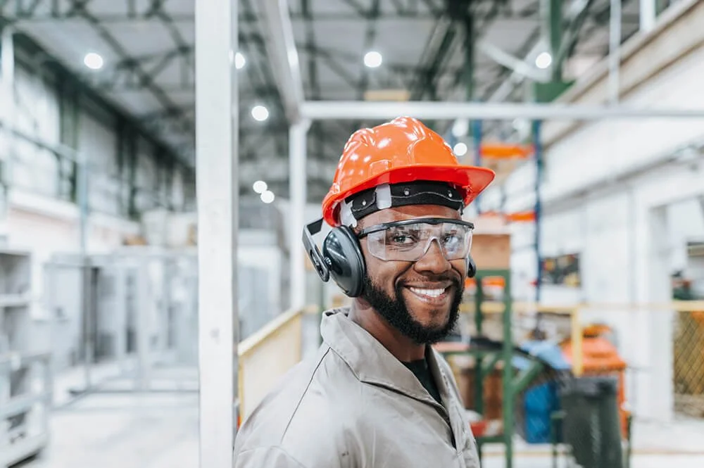 A worker wearing safety gear, including a hard hat, safety glasses, and ear protection, smiling in an industrial warehouse.