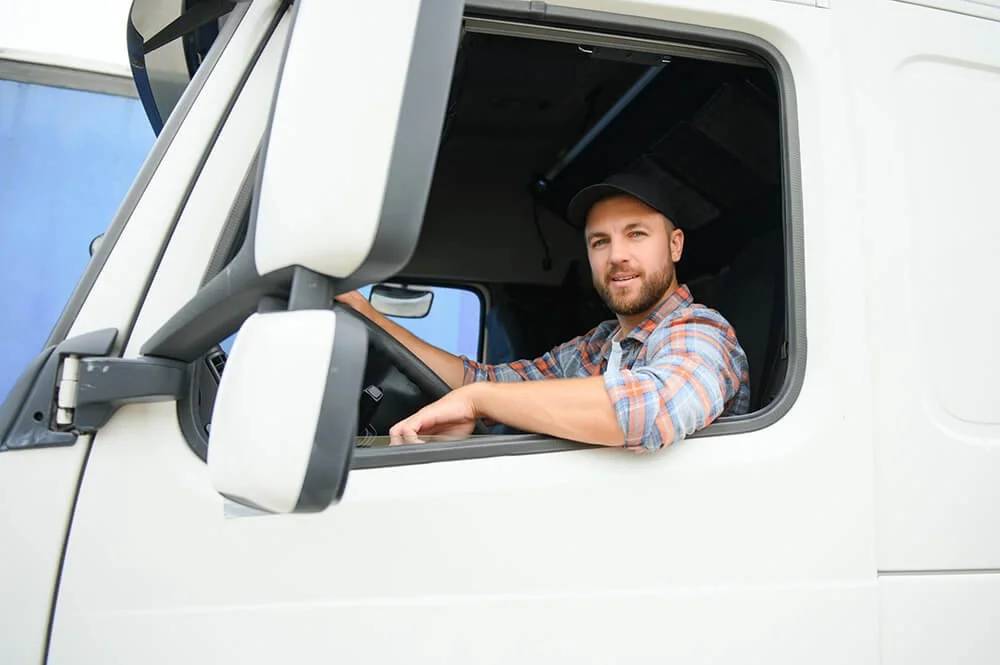 A man with a beard and plaid shirt sitting in the driver's seat of a white truck, looking out the window.