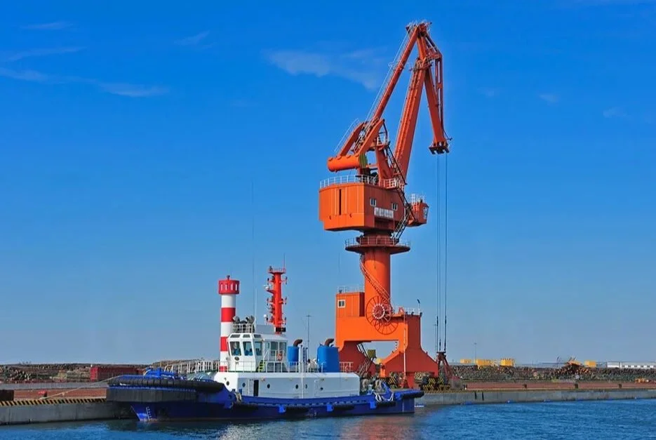 A large orange harbor crane on a dock near a white tugboat with red and white striped smokestacks.