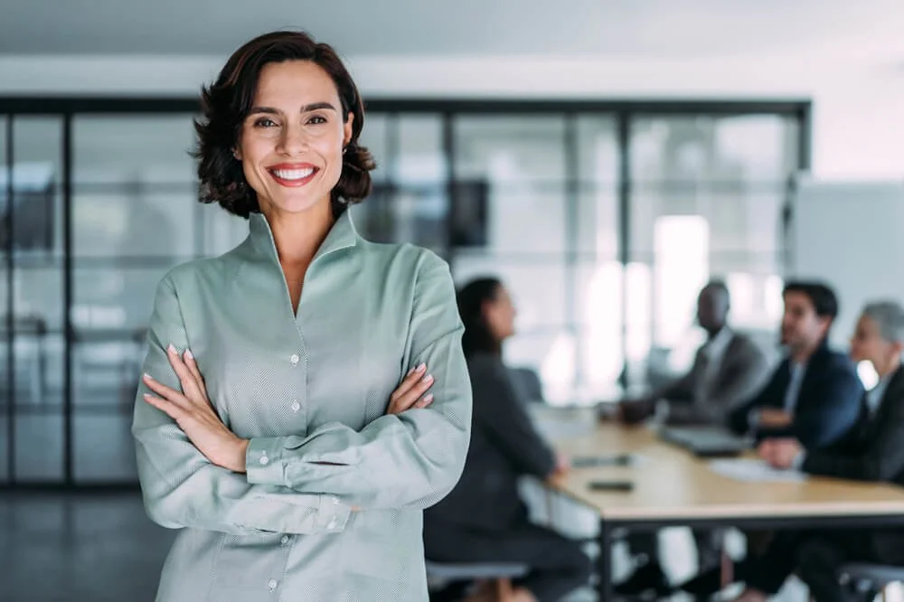 Businesswoman smiling with arms crossed in a corporate meeting room with colleagues in the background.