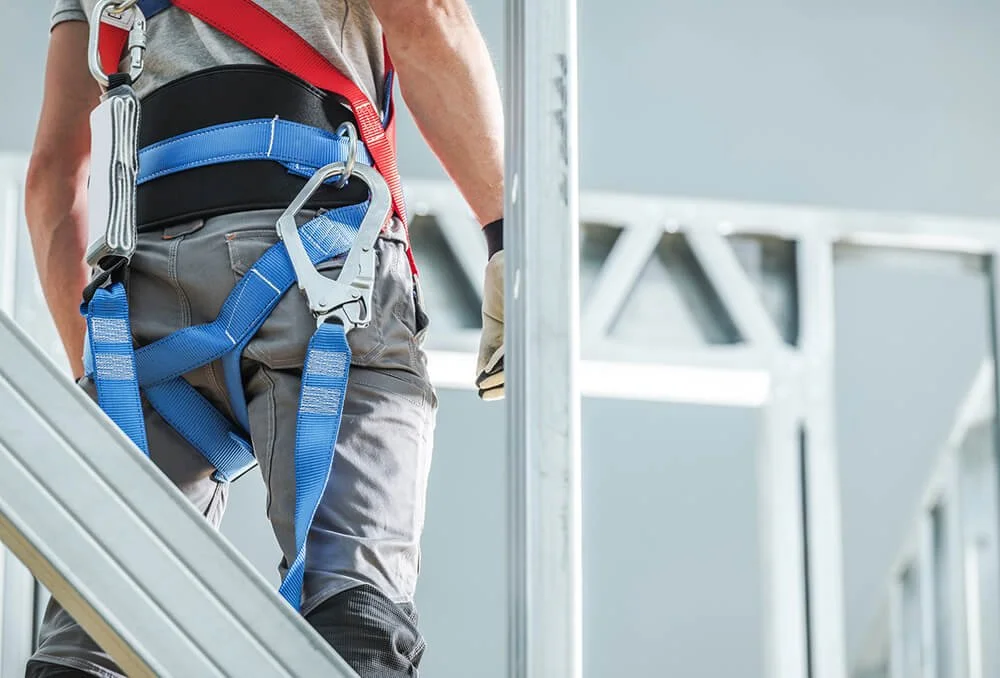 A construction worker wearing a safety harness with blue straps is working on a building framework.
