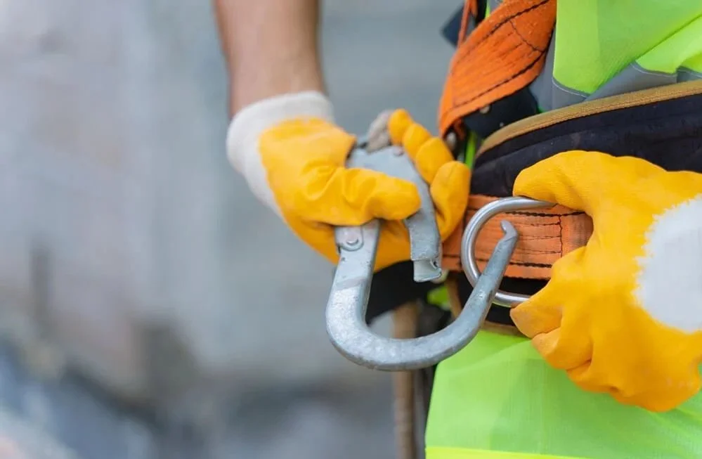 Close-up of a construction worker's hands fastening a safety harness with hooks and carabiners, wearing yellow gloves.