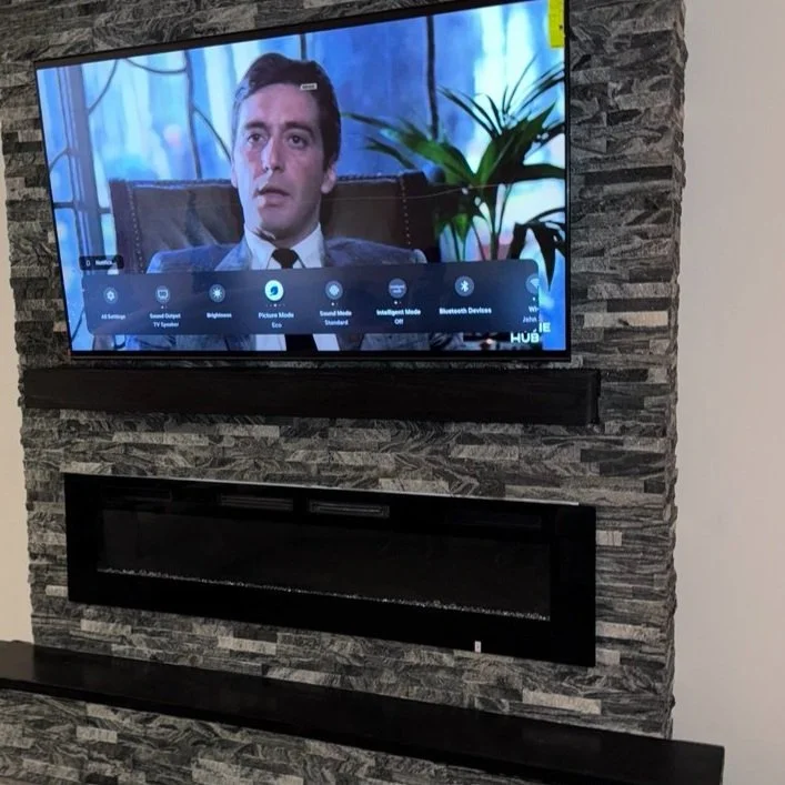 A flat-screen TV mounted on a stone wall, displaying a scene from a TV show or movie featuring a man in a suit, with a plant visible in the background.