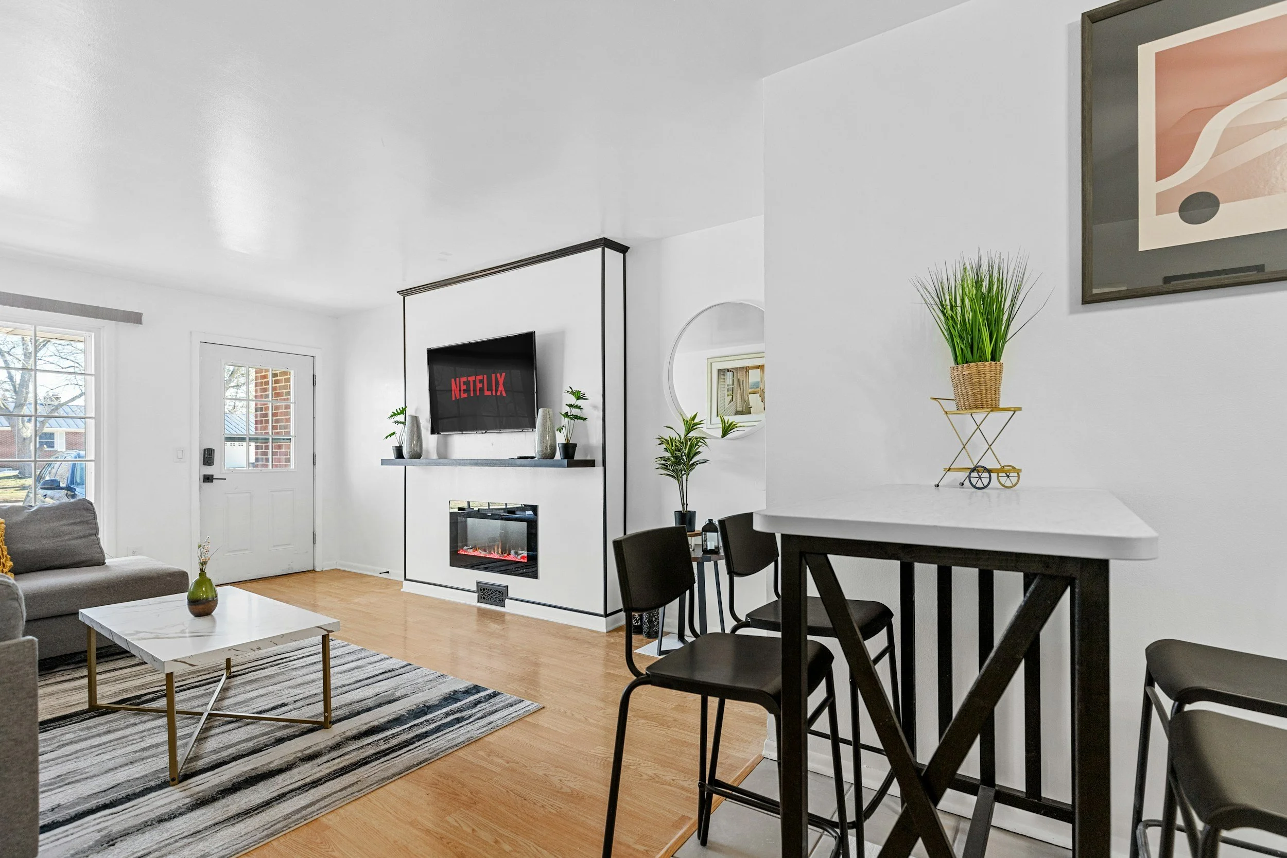 Living room with white walls, a gray couch, a black and white striped rug, a white marble coffee table, a wall-mounted television displaying Netflix, a small electric fireplace below it, and a dining area with black chairs and a white countertop with potted plants.