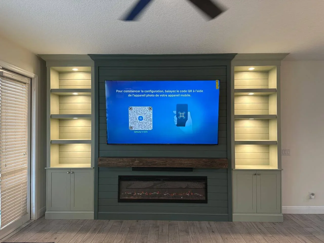 Green shiplap entertainment center in Santa Ana with a TV, electric fireplace, wood mantel, illuminated shelves, and cabinets.