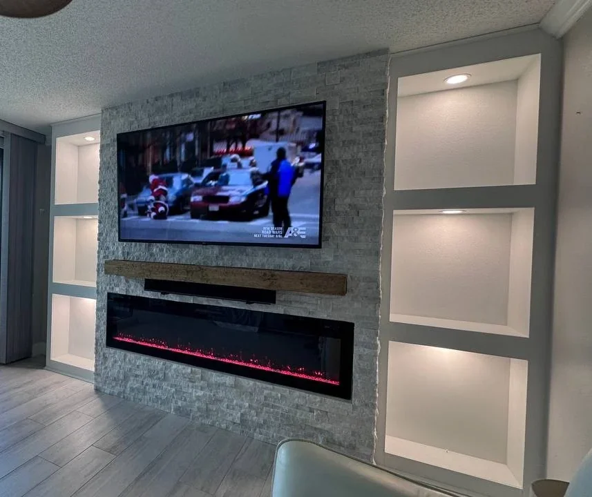 Gray stone media wall in Pasadena with a TV, electric fireplace, wood mantel, and six illuminated display niches.