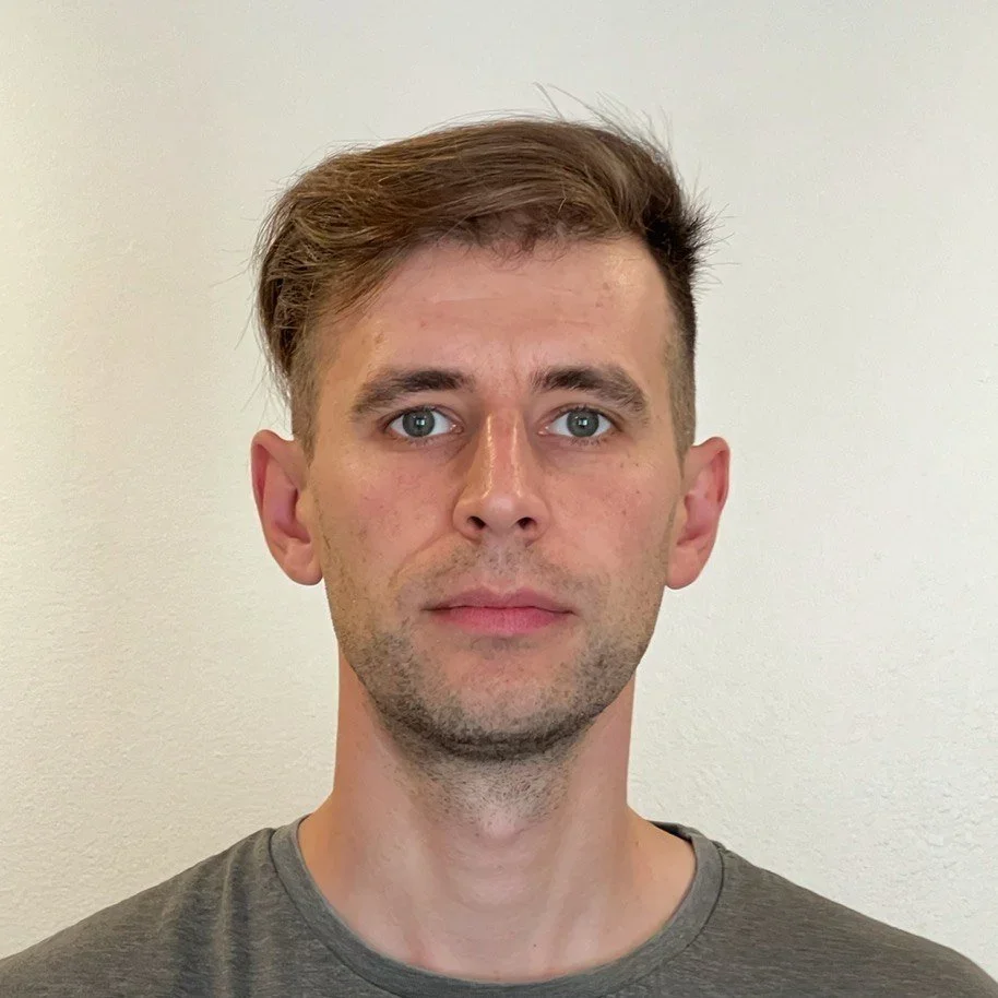 A young man with short brown hair, fair skin, and blue eyes standing in front of a plain light-colored wall.