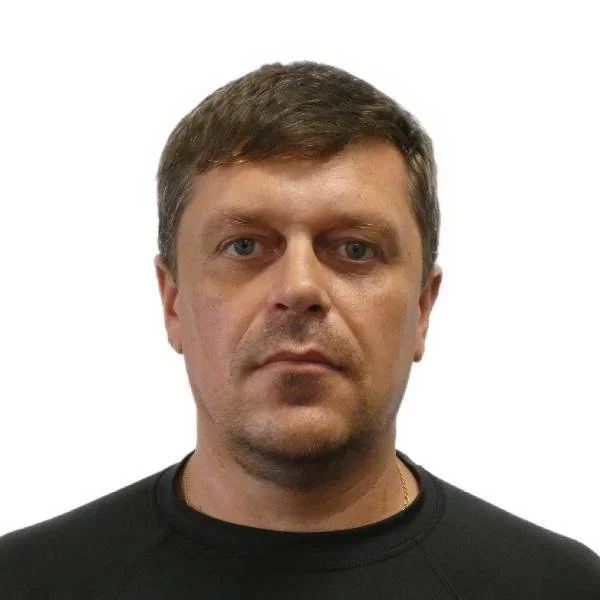 A portrait of a man with short brown hair, blue eyes, and light skin, wearing a black shirt, against a plain white background.