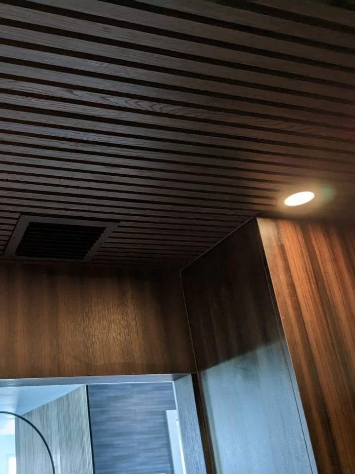 Ceiling with wooden slats, a round recessed light, and an air vent, part of an interior ceiling design.