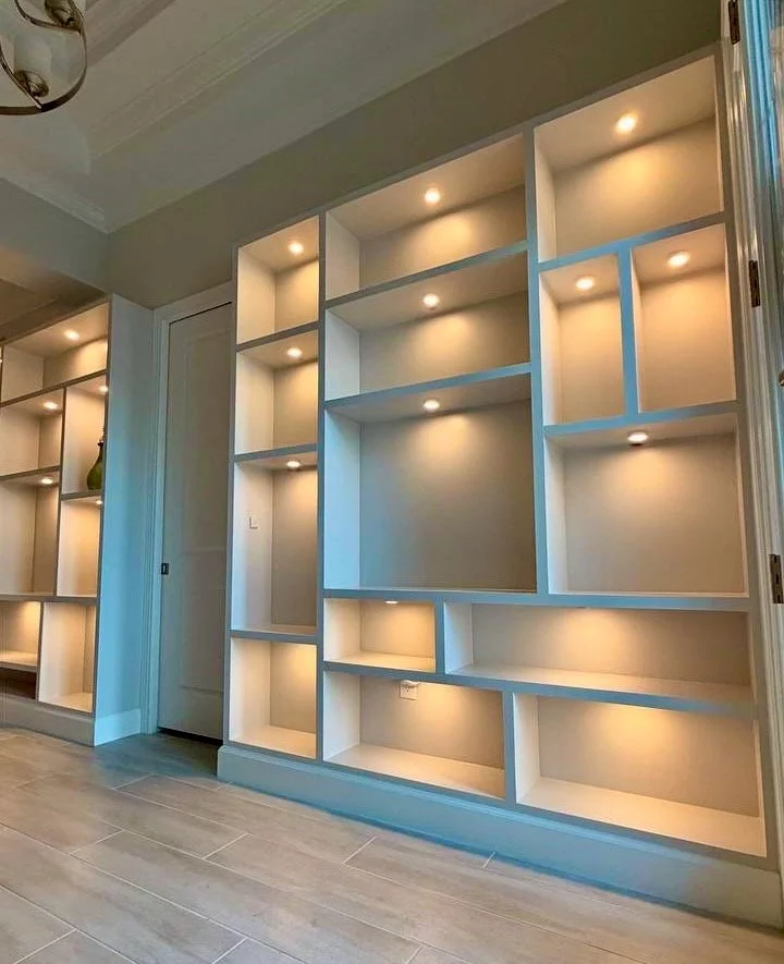 Angled view of a white built-in display wall with varied niche sizes, warm lighting, and matching base trim.