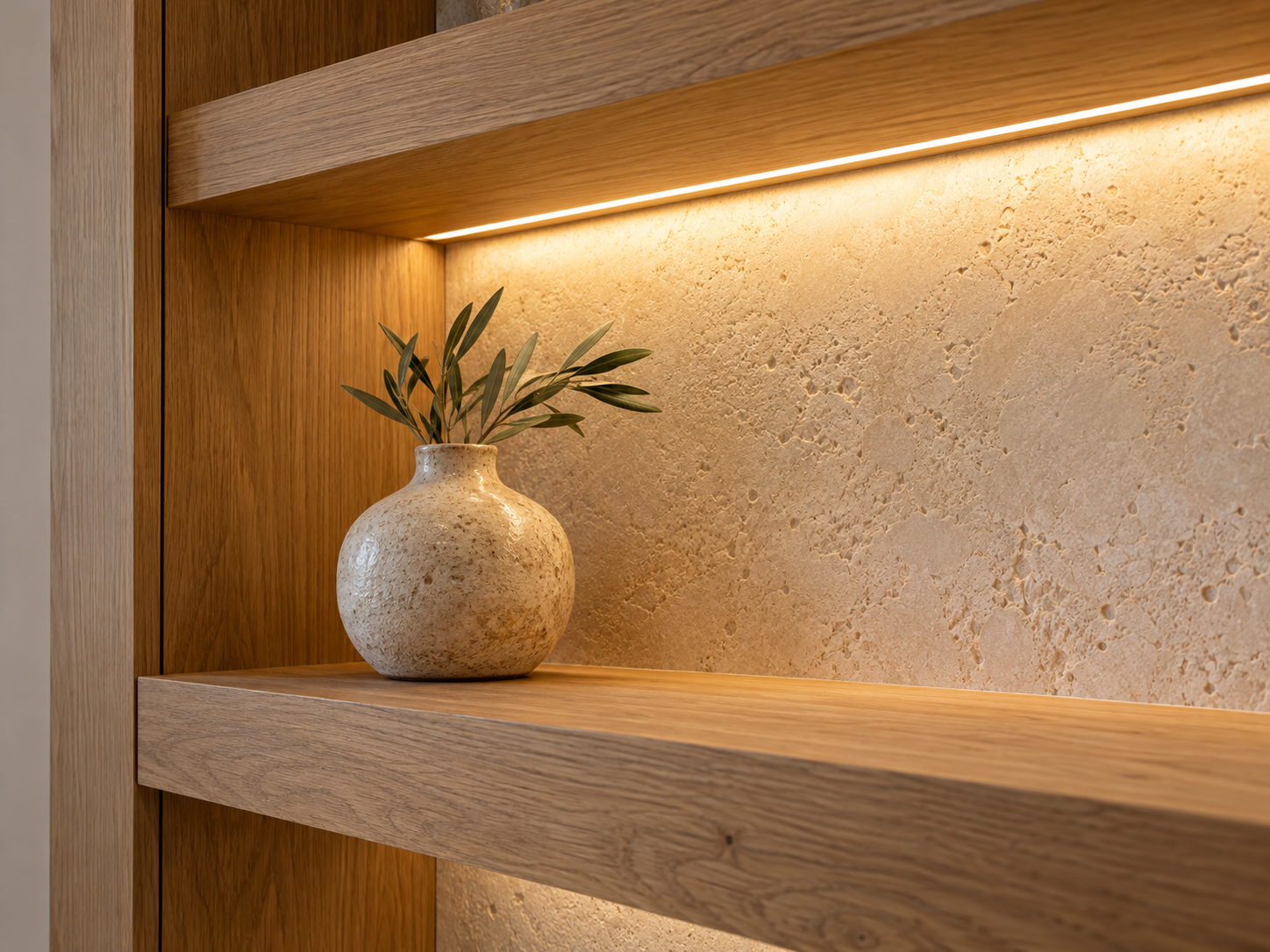 A beige textured ceramic vase with green leaves on a wooden shelf inside a built-in wooden alcove with a warm light strip above.