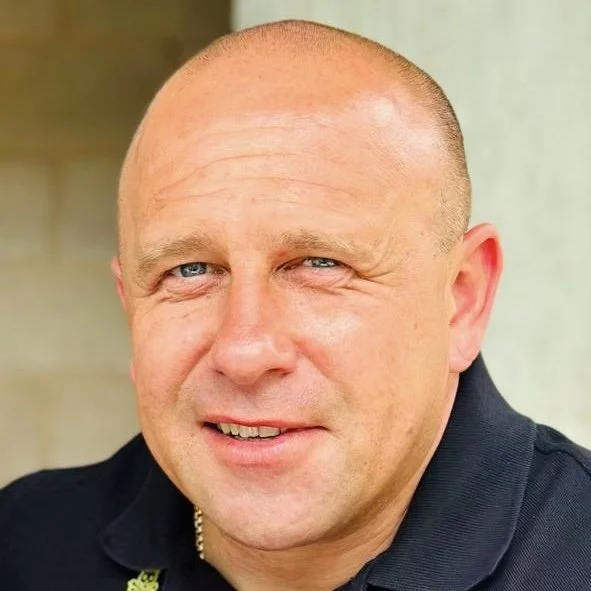 Close-up portrait of a middle-aged man with a shaved head, blue eyes, and a slight smile, wearing a dark shirt.