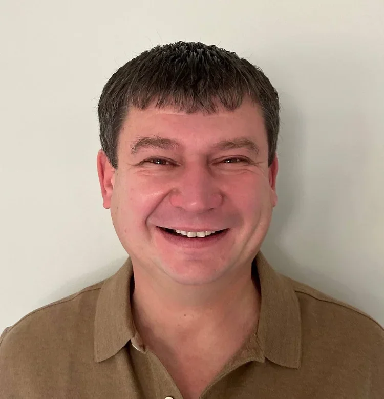 A smiling man with short dark hair wearing a brown polo shirt, standing in front of a plain white wall.