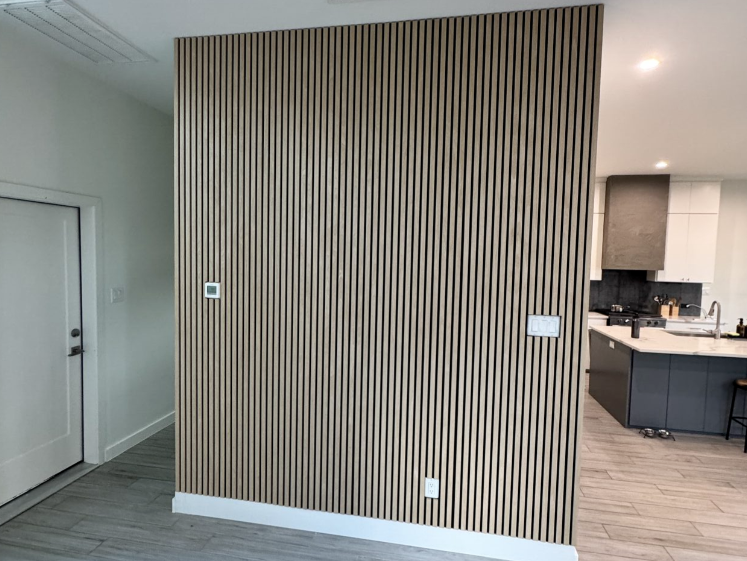Full-height wood slat accent wall in a Dallas home, with light wood vertical slats and dark backing beside an open kitchen.