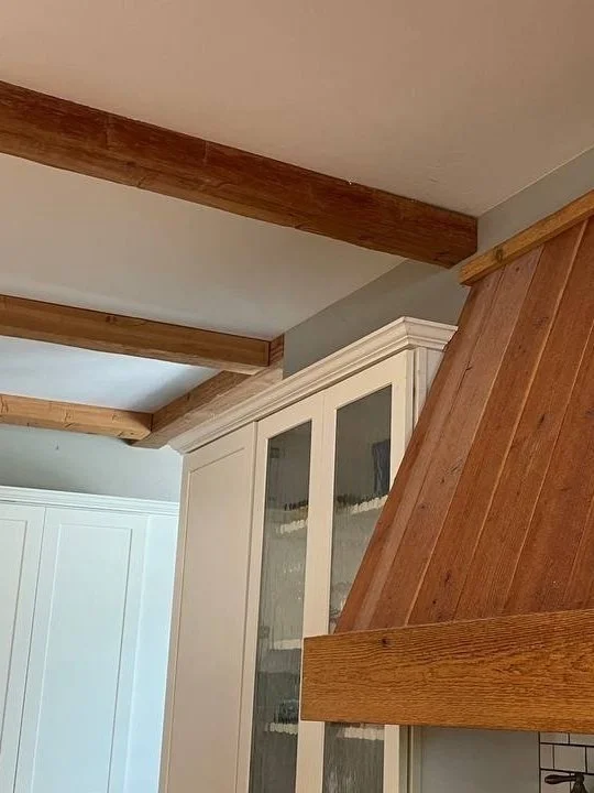 View of kitchen ceiling with wooden beams, a white cabinet with glass doors, and part of a wooden kitchen hood or range hood.