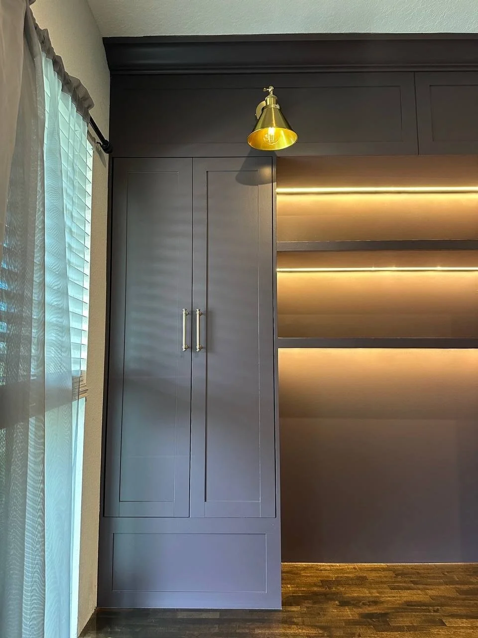 Tall painted office cabinet beside warm illuminated shelves, with a brass-toned sconce and wood desktop.