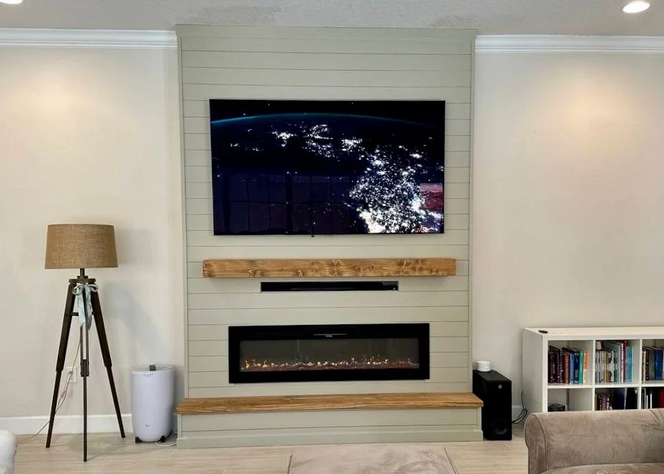 Living room with a mounted TV showing outer space; fireplace below the TV; wooden mantel and shelf; tall floor lamp to the left; bookshelf on the right.