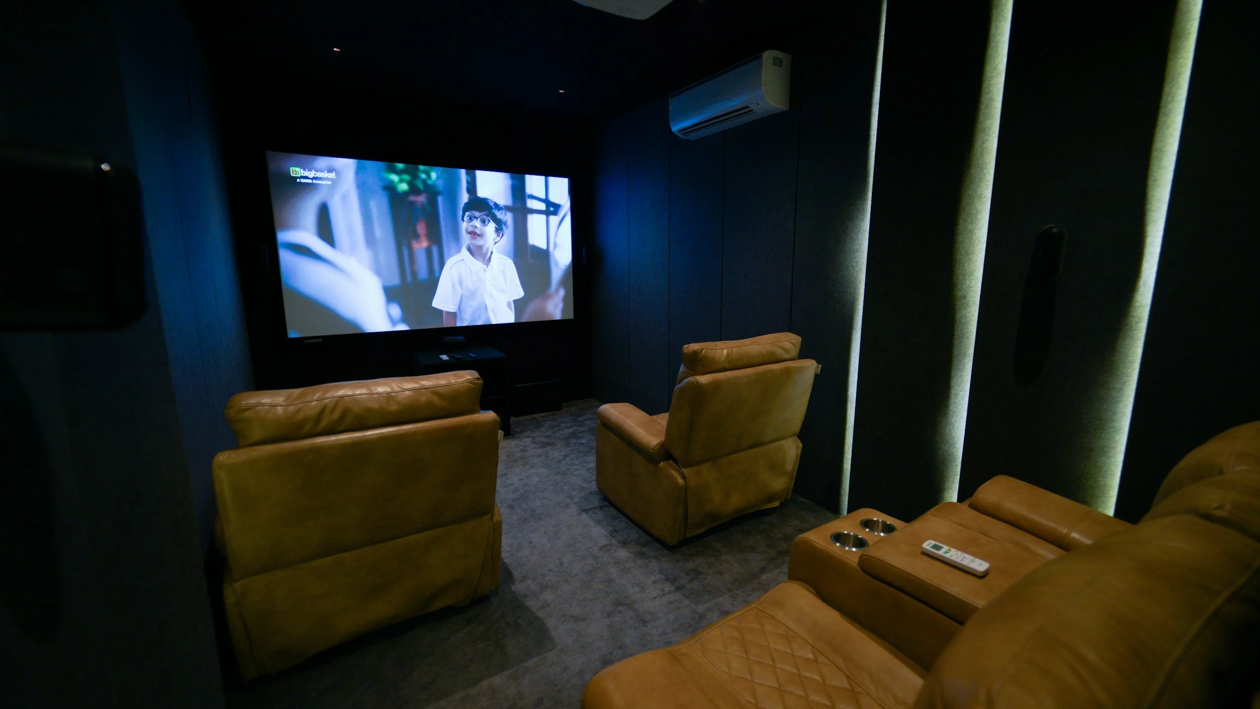 A private home theater with a large television screen showing a young boy in eyeglasses. There are brown leather recliner chairs and a remote control on the armrest.