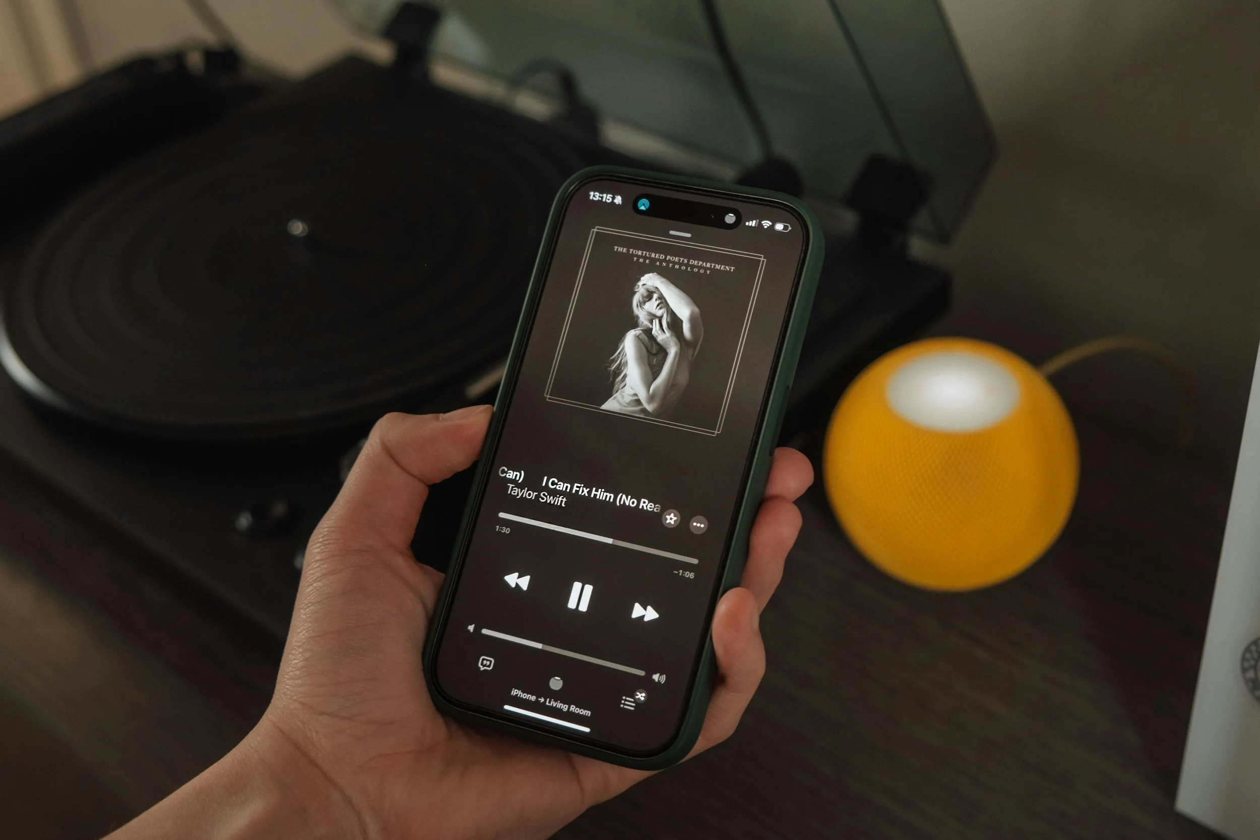 Person holding a smartphone playing a song titled 'I Can Fix Him (No Reason)' by Taylor Swift, with a turntable and yellow smart speaker on a dark table in the background.