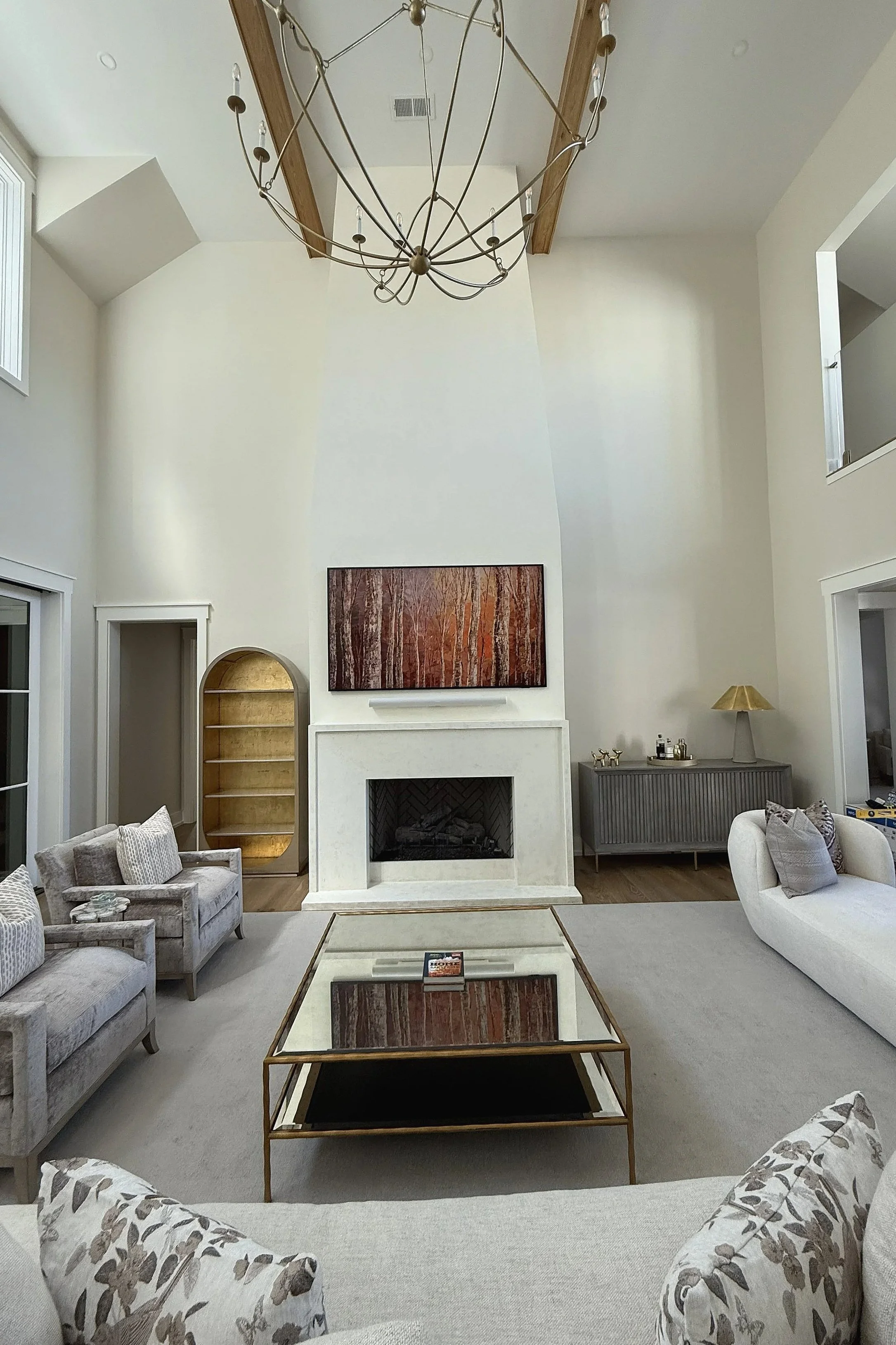 A living room with high ceilings, featuring a modern fireplace, a large flat-screen TV, a chandelier, and various seating options including upholstered couches and armchairs. There is a glass coffee table and decorative items such as pillows, lamps, 