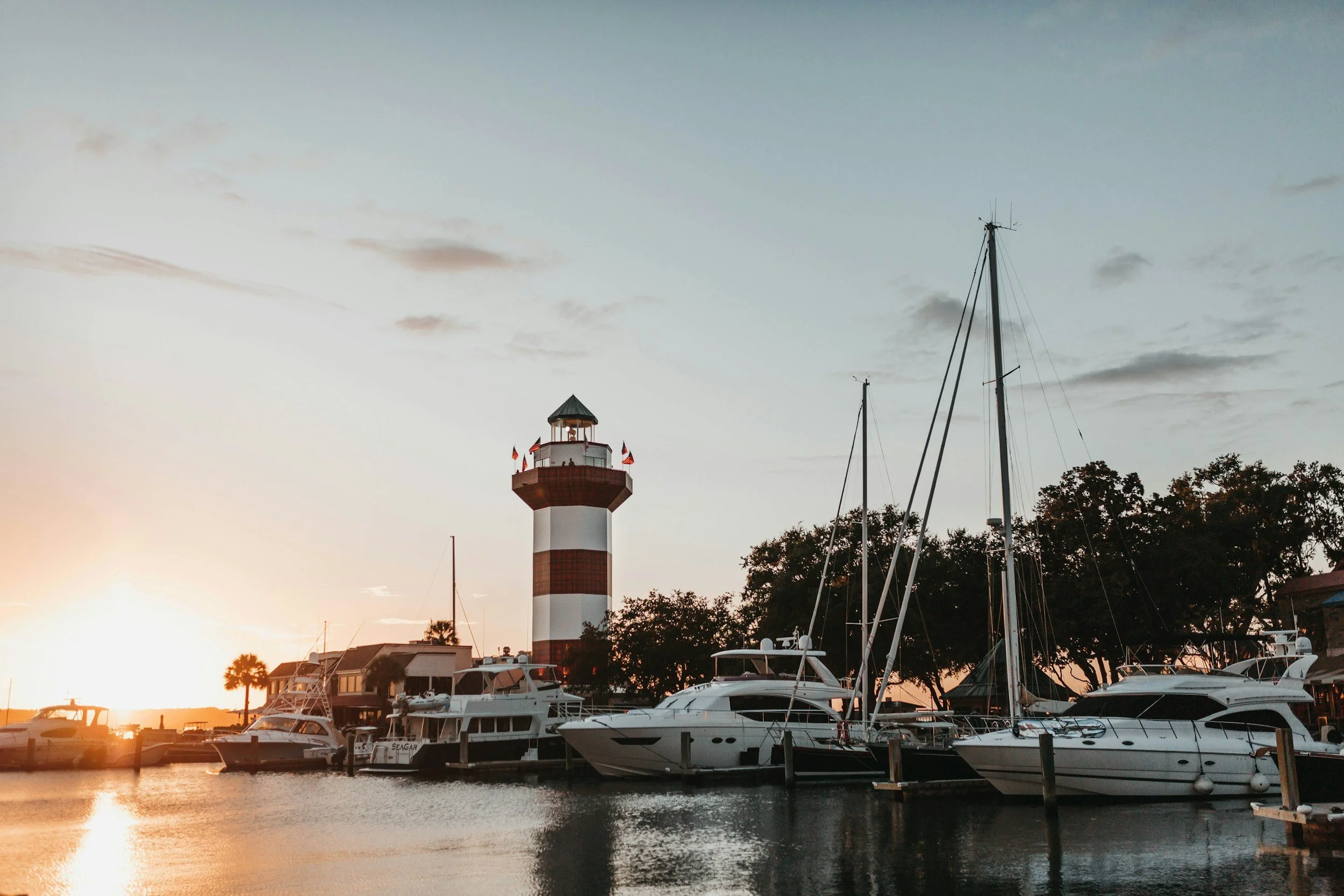 Boats docked at a marina with a striped lighthouse in the background during sunset.