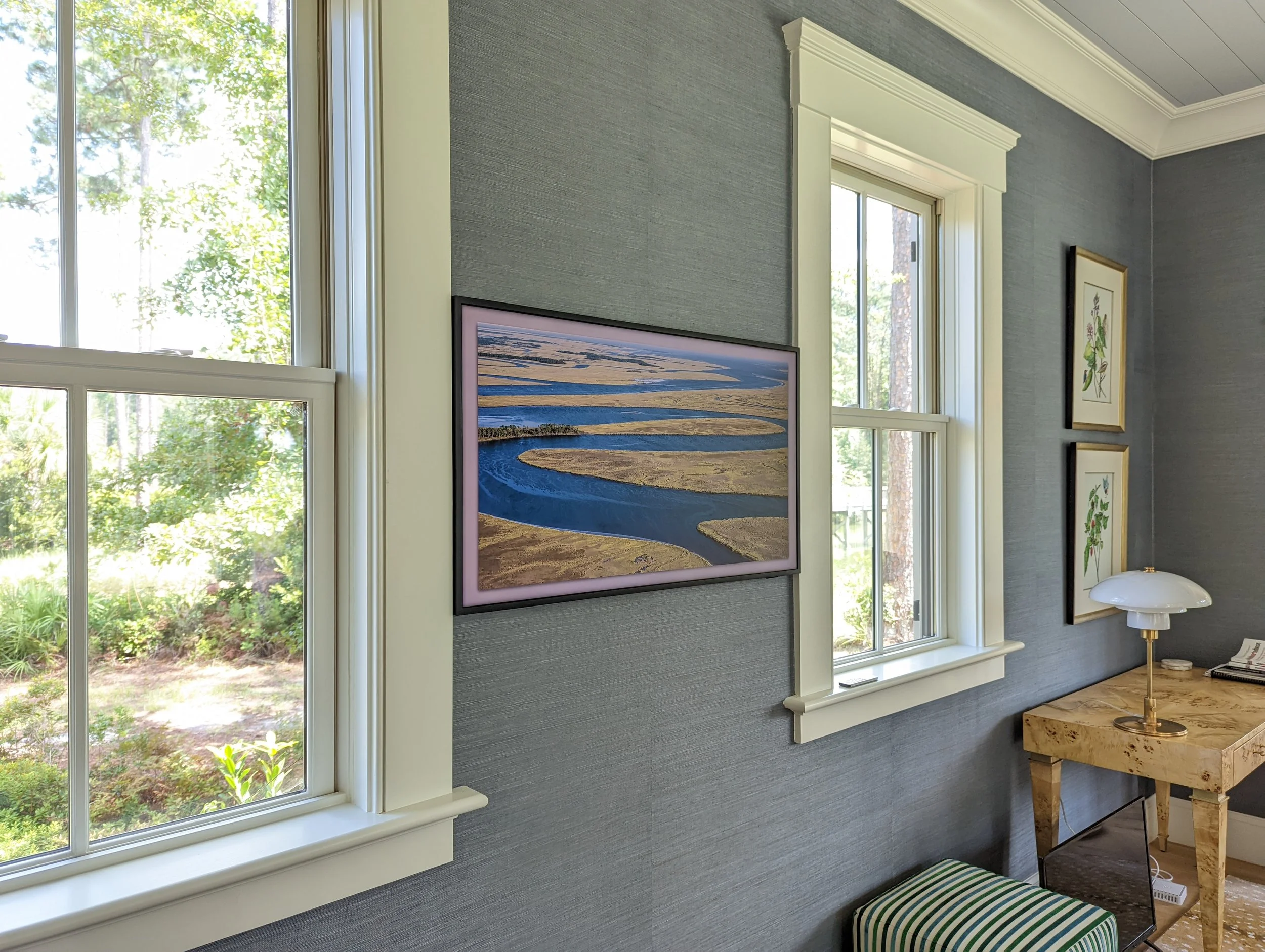 Interior of a room with blue textured walls, two windows with white trim, a wall-mounted landscape photograph of a river and wetlands, and two framed botanical prints. There's a wooden desk with a white lamp and a small striped ottoman.