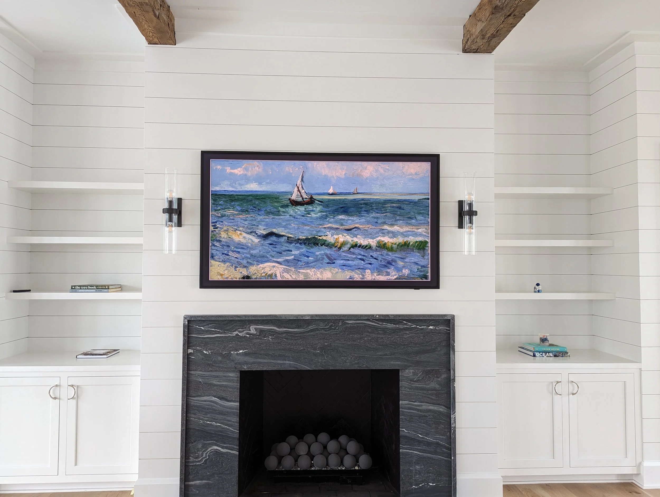 Living room with a fireplace, white shiplap walls, and built-in shelves on either side. A framed painting of a sailboat on the ocean is above the fireplace. Two wall-mounted candle sconces flank the painting, and shelves hold books and small decor it
