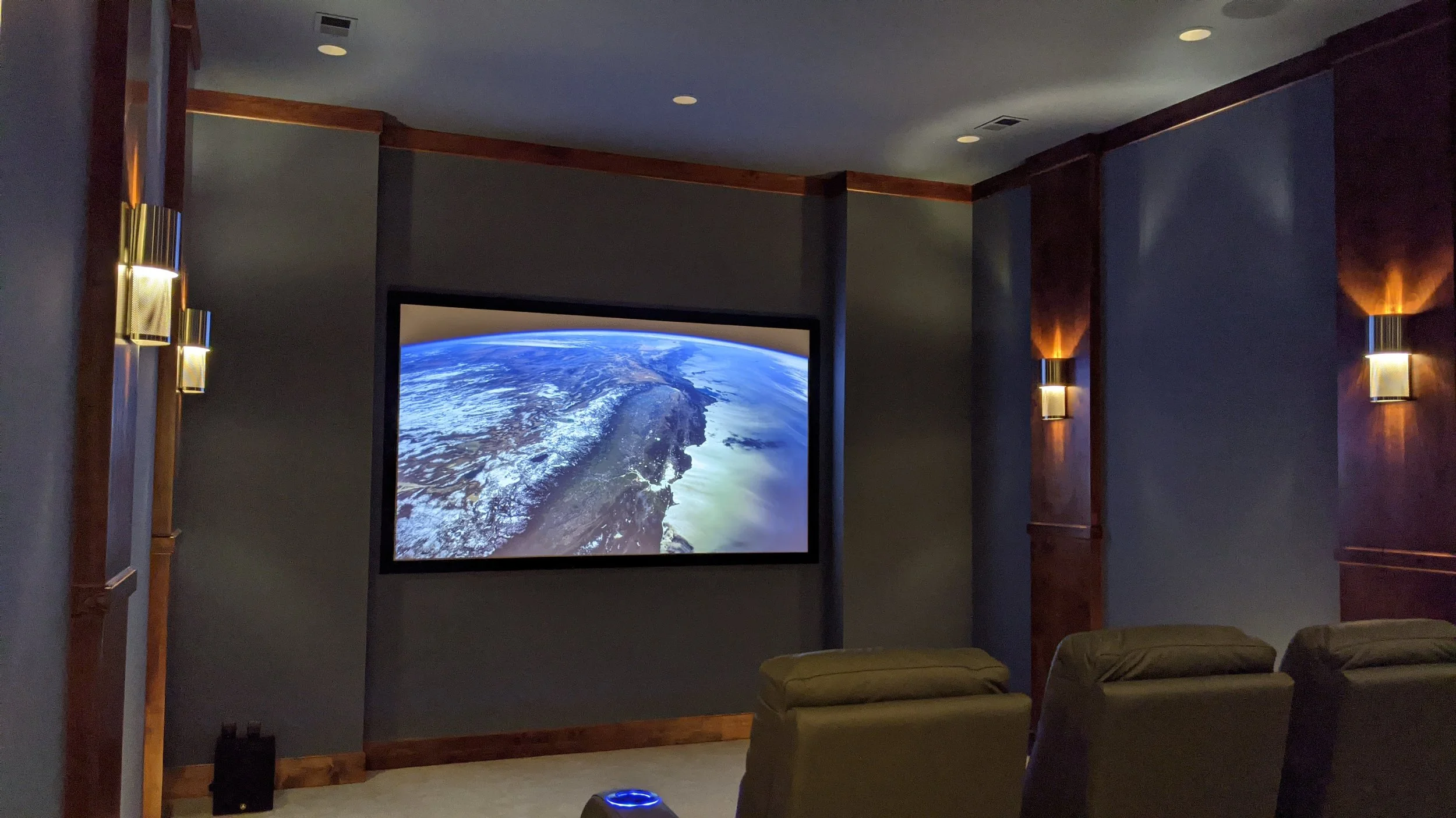 Home theater room with a large screen displaying an aerial view of a coast and ocean, soft lighting, blue walls, and three theater chairs facing the screen.