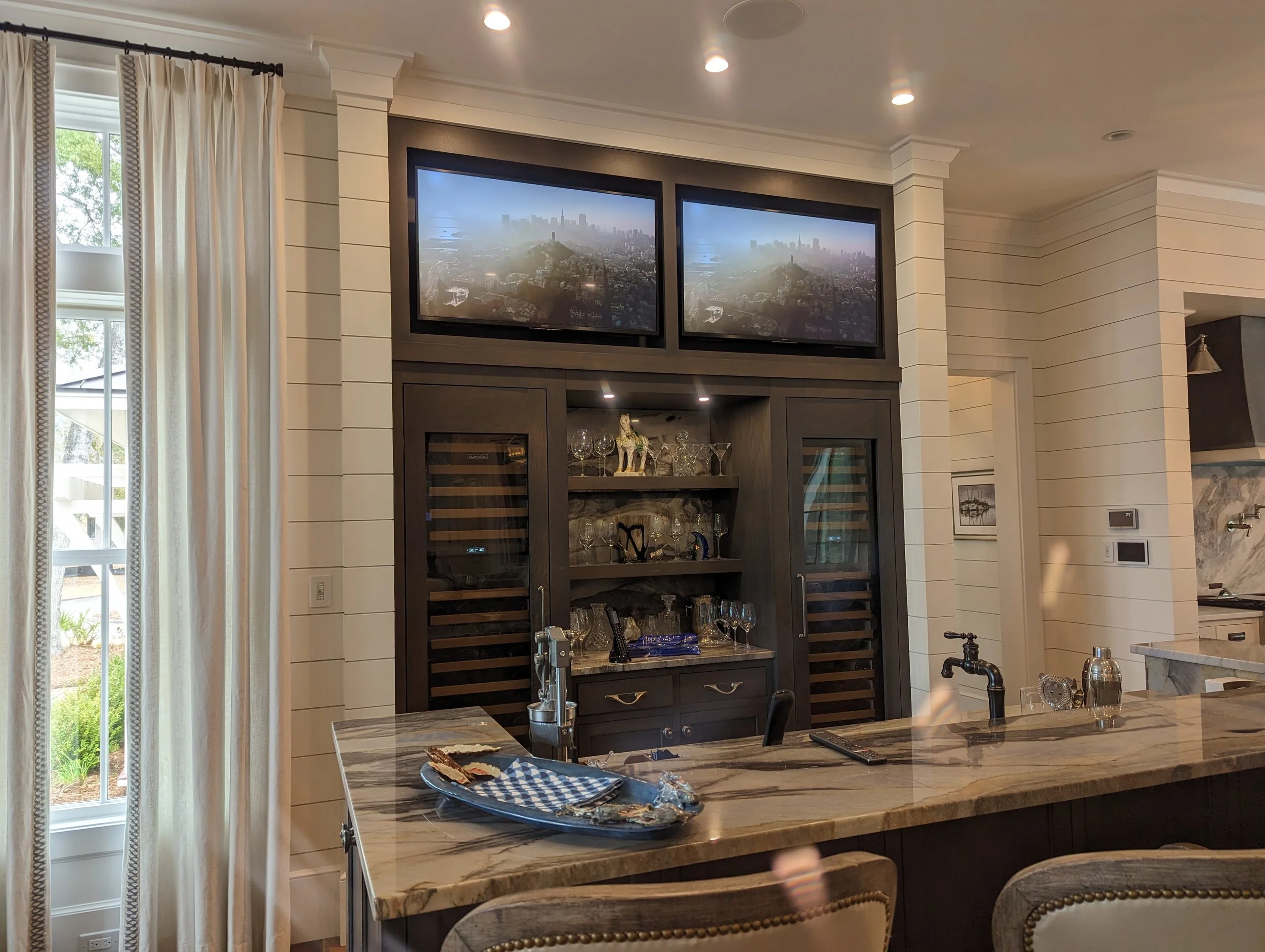 Interior of a modern kitchen with a marble island, a built-in cabinet with glass doors displaying glassware, and two TVs mounted on the wall showing an aerial city view.