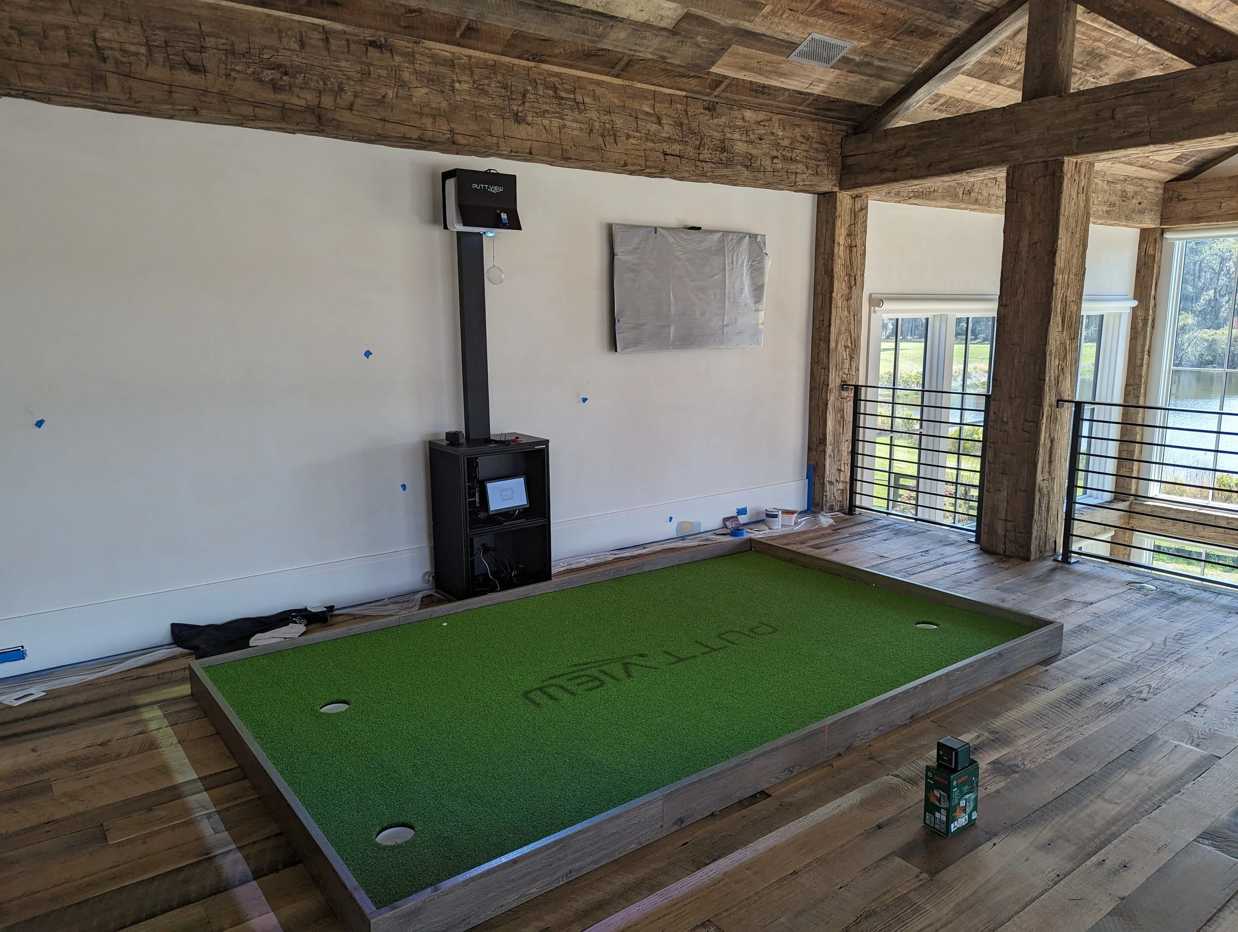 Interior of a room with a raised putting green, large wooden beams in the ceiling, windows showing an outdoor view of grass and trees, construction materials along the wall, and a black equipment stand with a screen and some equipment on top.