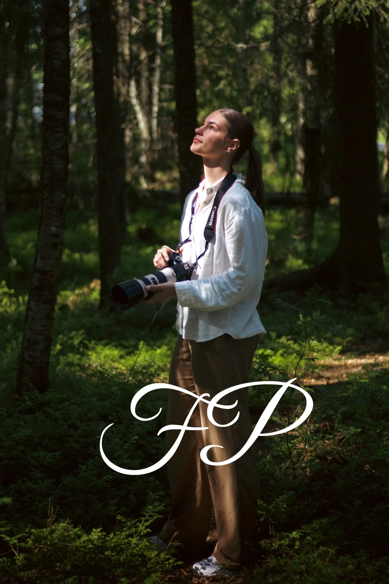 A woman holding a camera with a long lens in a forest, dressed in a white shirt and brown pants, looking upward with sunlight filtering through the trees.