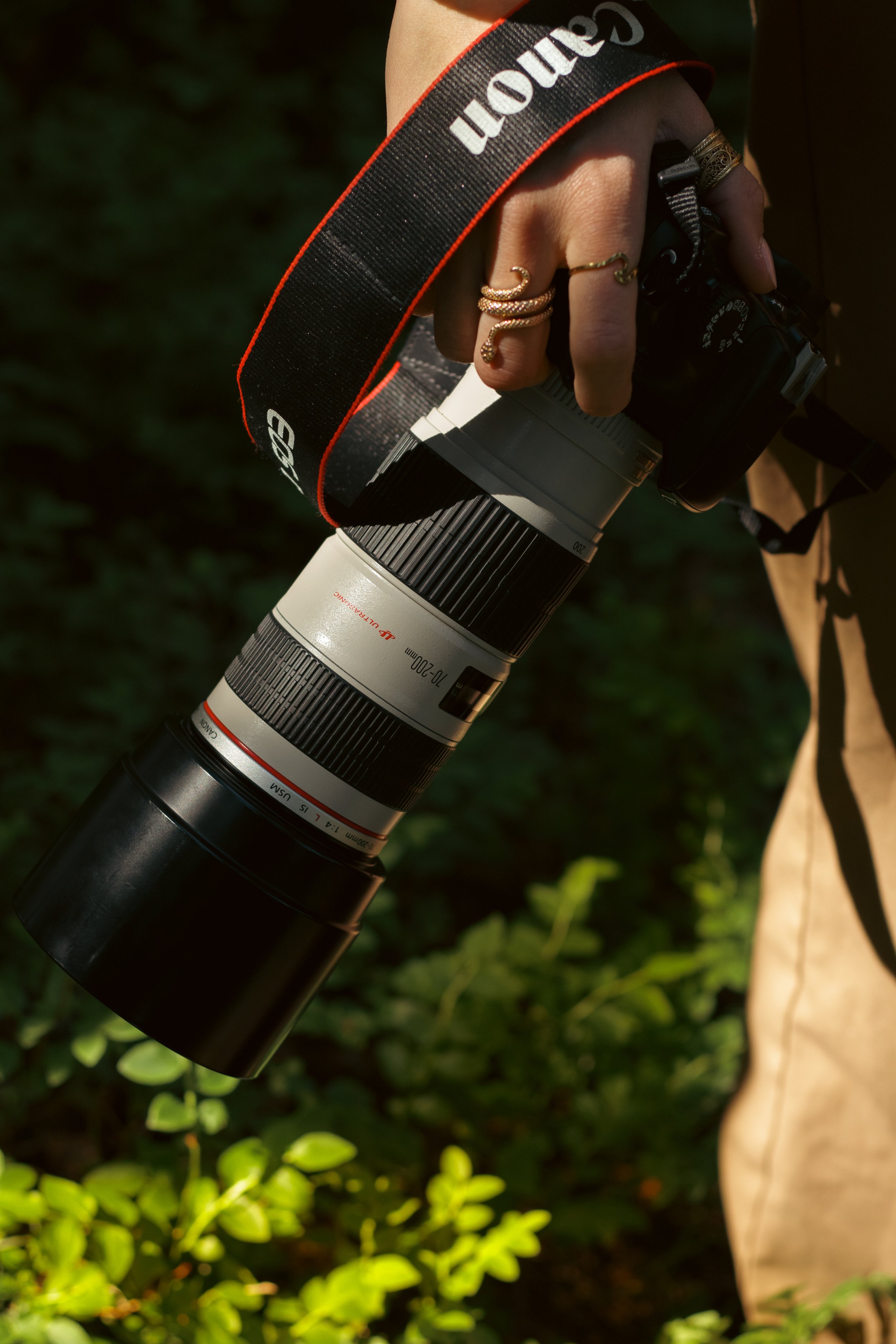 Close-up of a person holding a Canon camera with a large zoom lens attached, standing outdoors among green foliage, wearing rings and a watch.