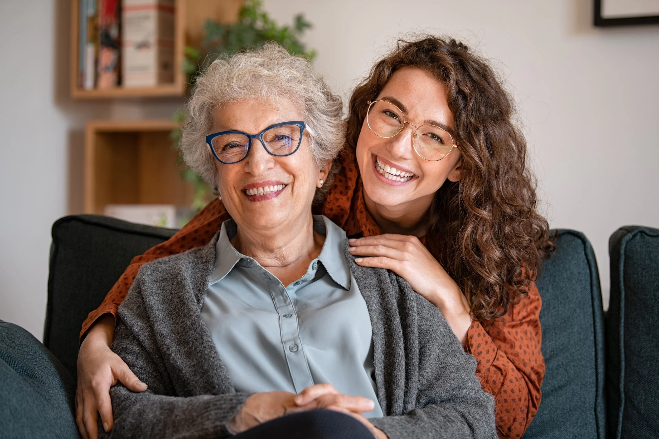 Two smiling women, one elderly with gray hair and glasses, and one young woman with curly hair and glasses, sitting on a couch in a living room, embracing each other.