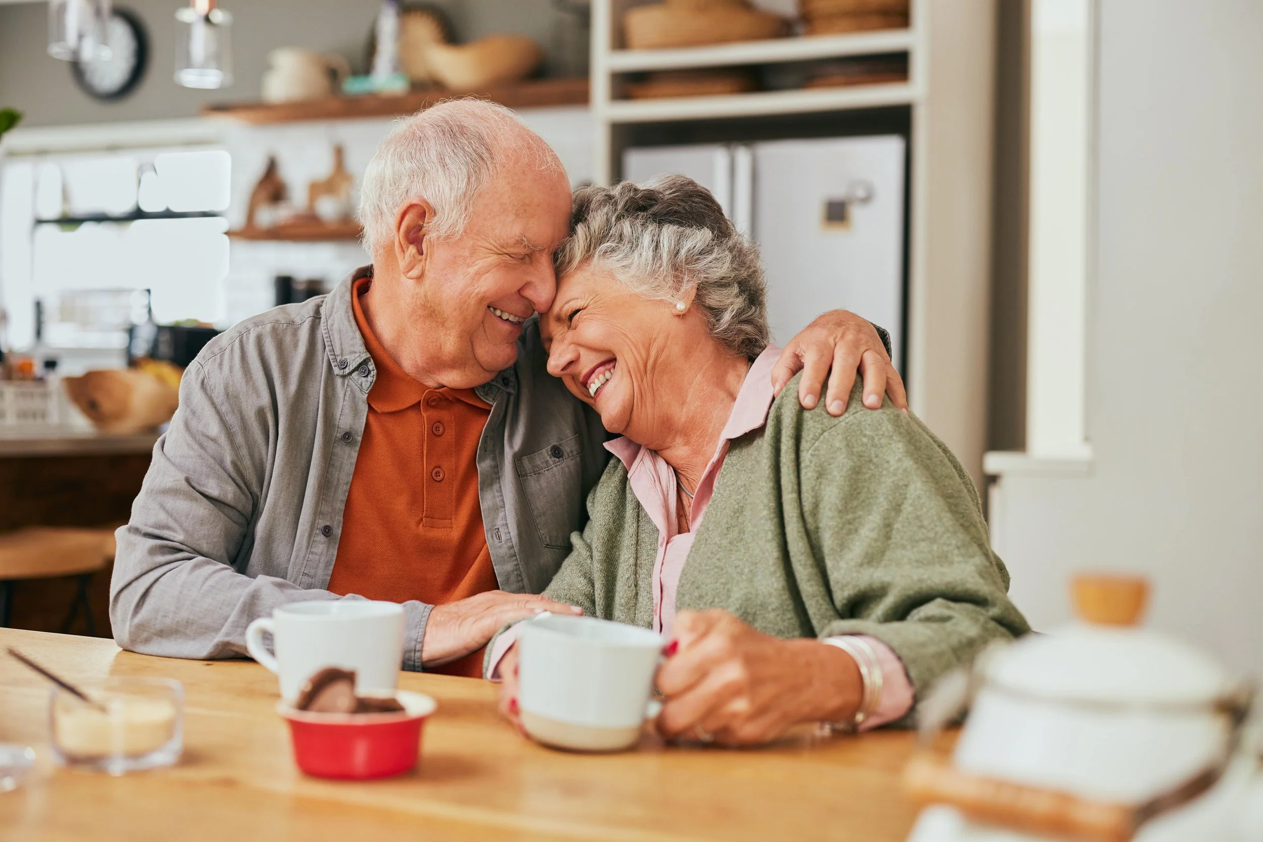 An elderly couple sitting at a kitchen table, smiling and leaning their foreheads together, enjoying each other's company.