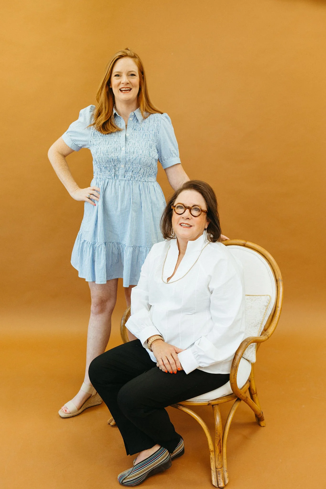 Two women posing against a brown background, one standing and one seated on a wicker chair. The seated woman wears glasses, a white blouse, black pants, and striped shoes. The standing woman wears a light blue and white striped dress and beige shoes.