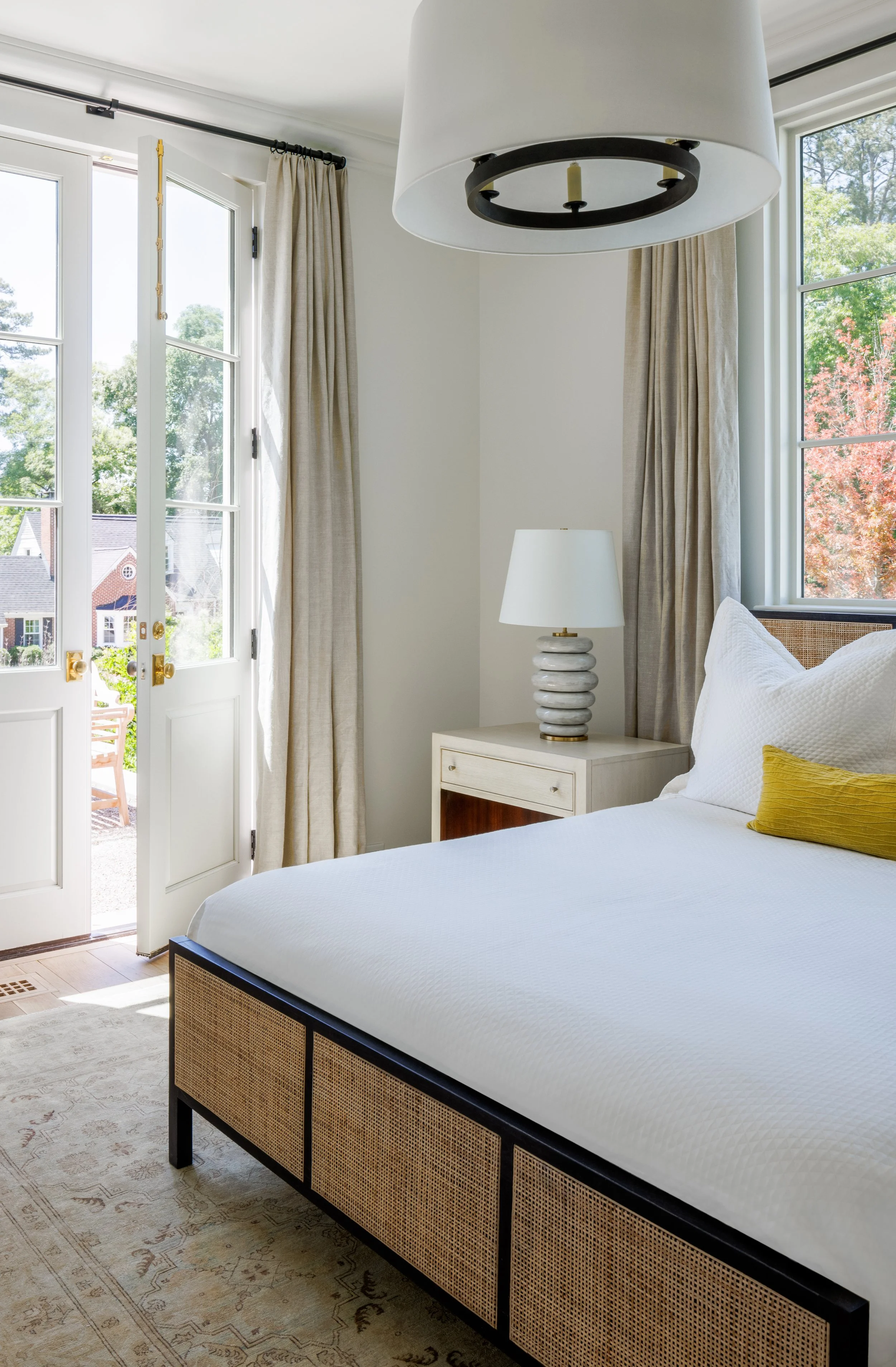 A bedroom with a large bed, white bedding, a bedside table with a white lamp, beige curtains, and large windows showing trees and sunlight outside.