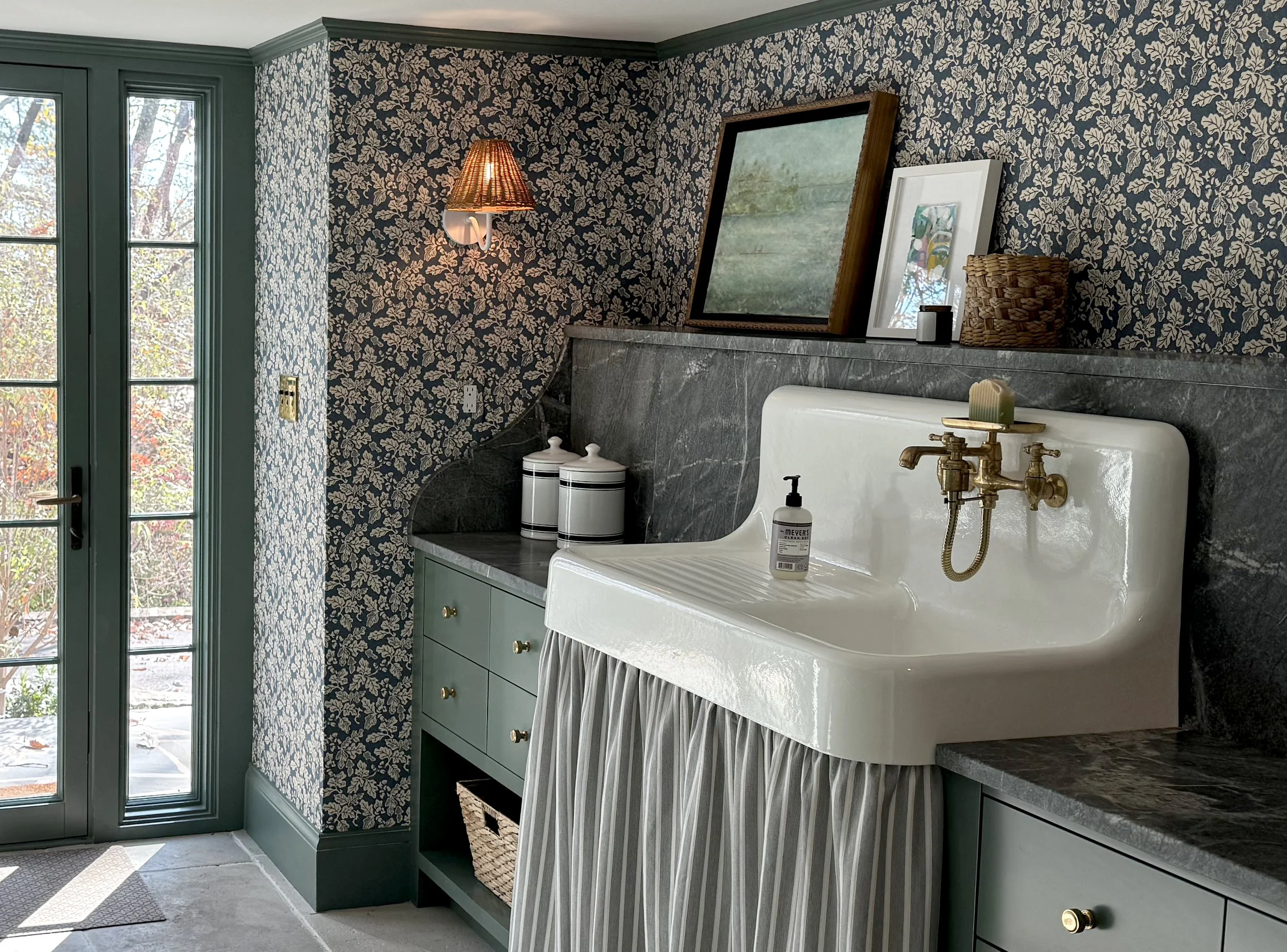Interior of a vintage bathroom with a green vanity, a white sink, and brass fixtures. The wall behind features dark wallpaper with a cream floral pattern. A large mirror with a wooden frame leans against the wall, and a wicker basket and framed artwo