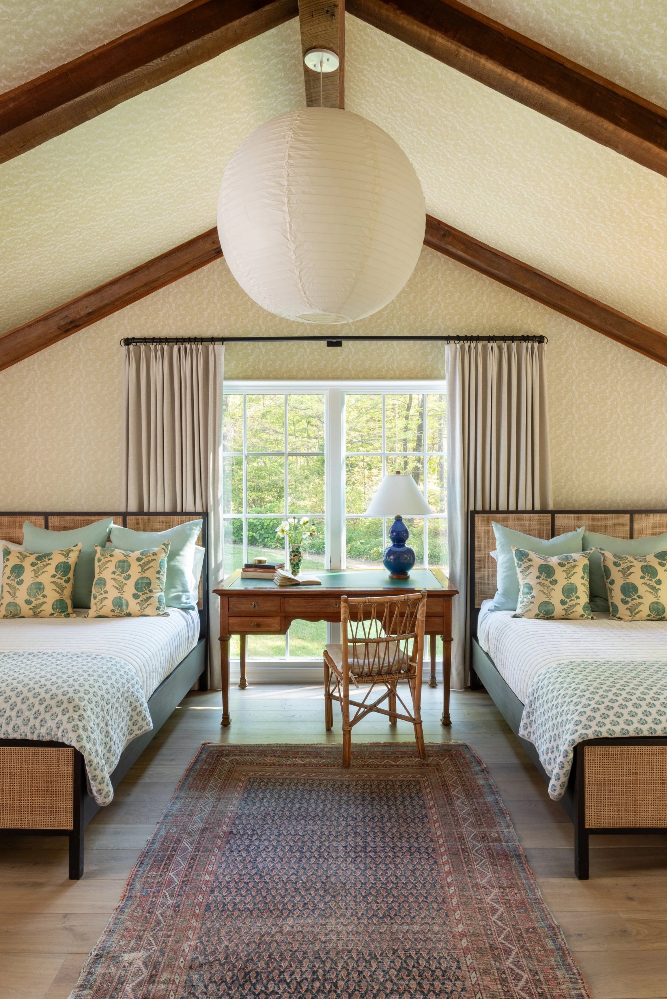 A bedroom featuring two beds with matching blue and white patterned bedding and green pillows, a central wooden desk with a blue lamp, a window with white curtains, a small table with a vase and books, a chair, and a large white paper lantern hanging