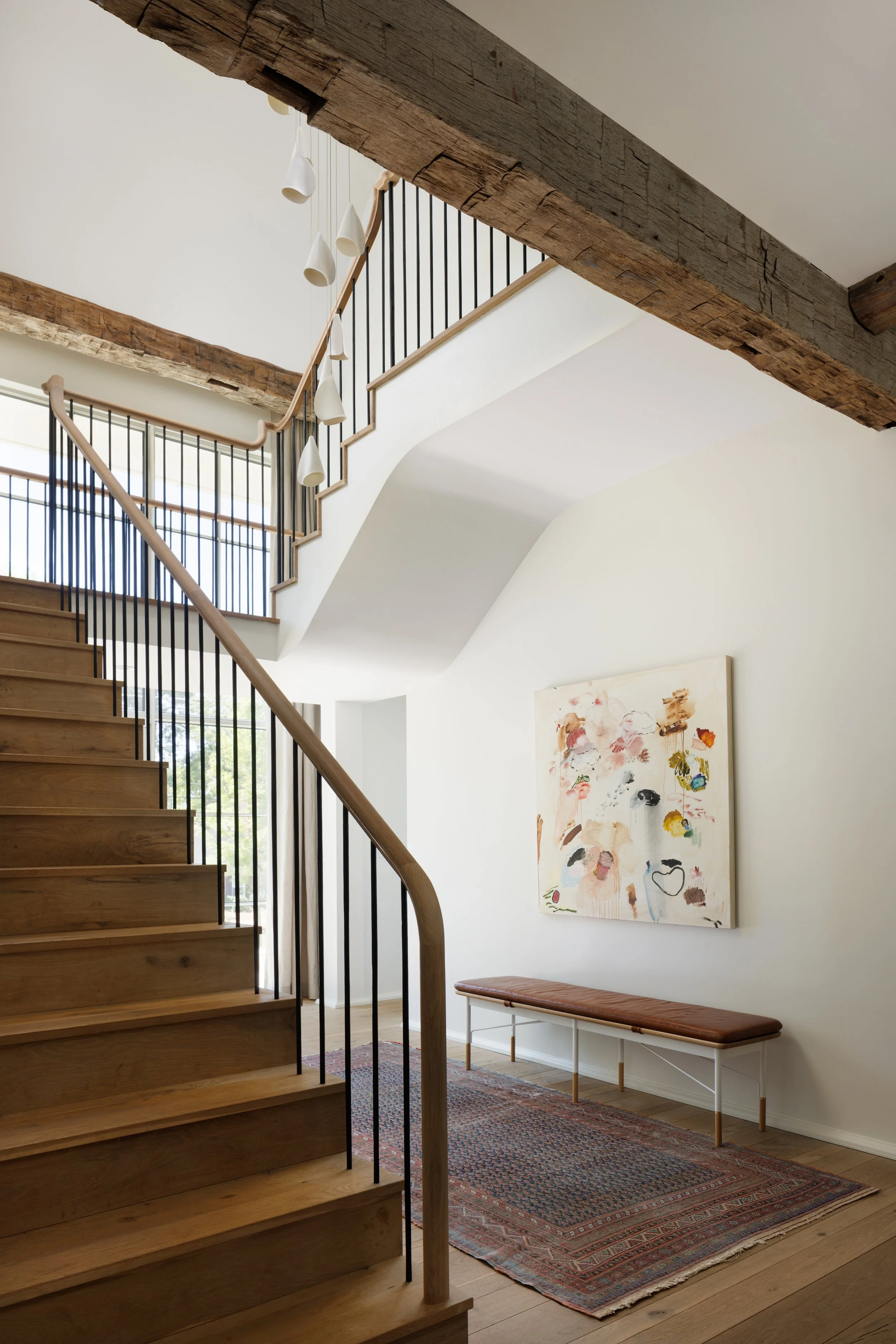 Wooden staircase with black metal railing leading up to a second floor. A white wall with a colorful abstract painting and a wooden bench on a patterned rug. Modern pendant lights hanging from the ceiling. Large window in the background letting in na