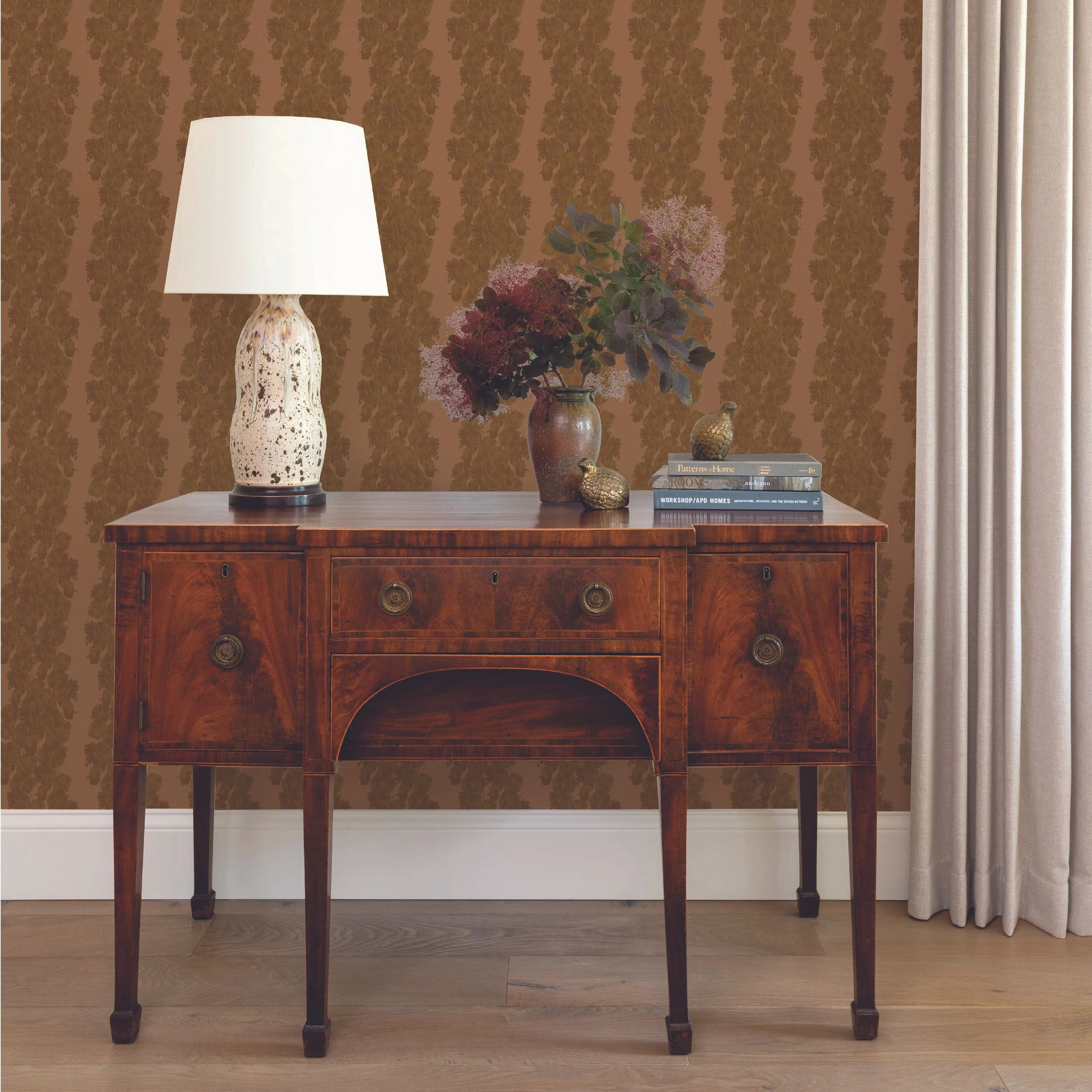 A wooden table with a decorative ceramic lamp, a vase with dried flowers, two small decorative figurines, and a stack of books, set against a brown wallpapered wall and beige curtains.