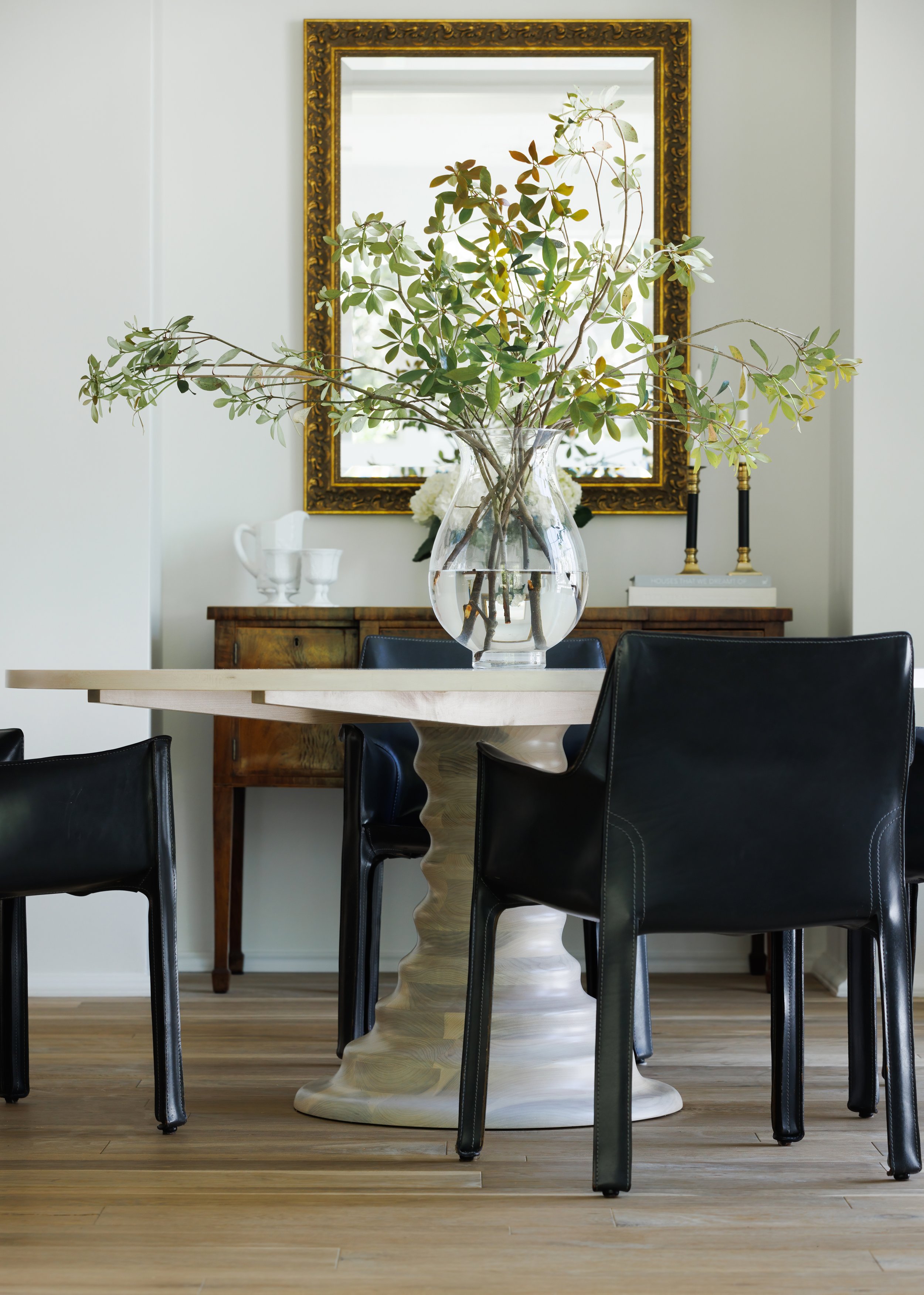 A modern dining room with a round, curly carved wooden table, a large clear glass vase with green leafy branches, a wooden sideboard with white dishes, a gold-framed mirror, and black chairs with leather upholstery around the table. A pair of black c