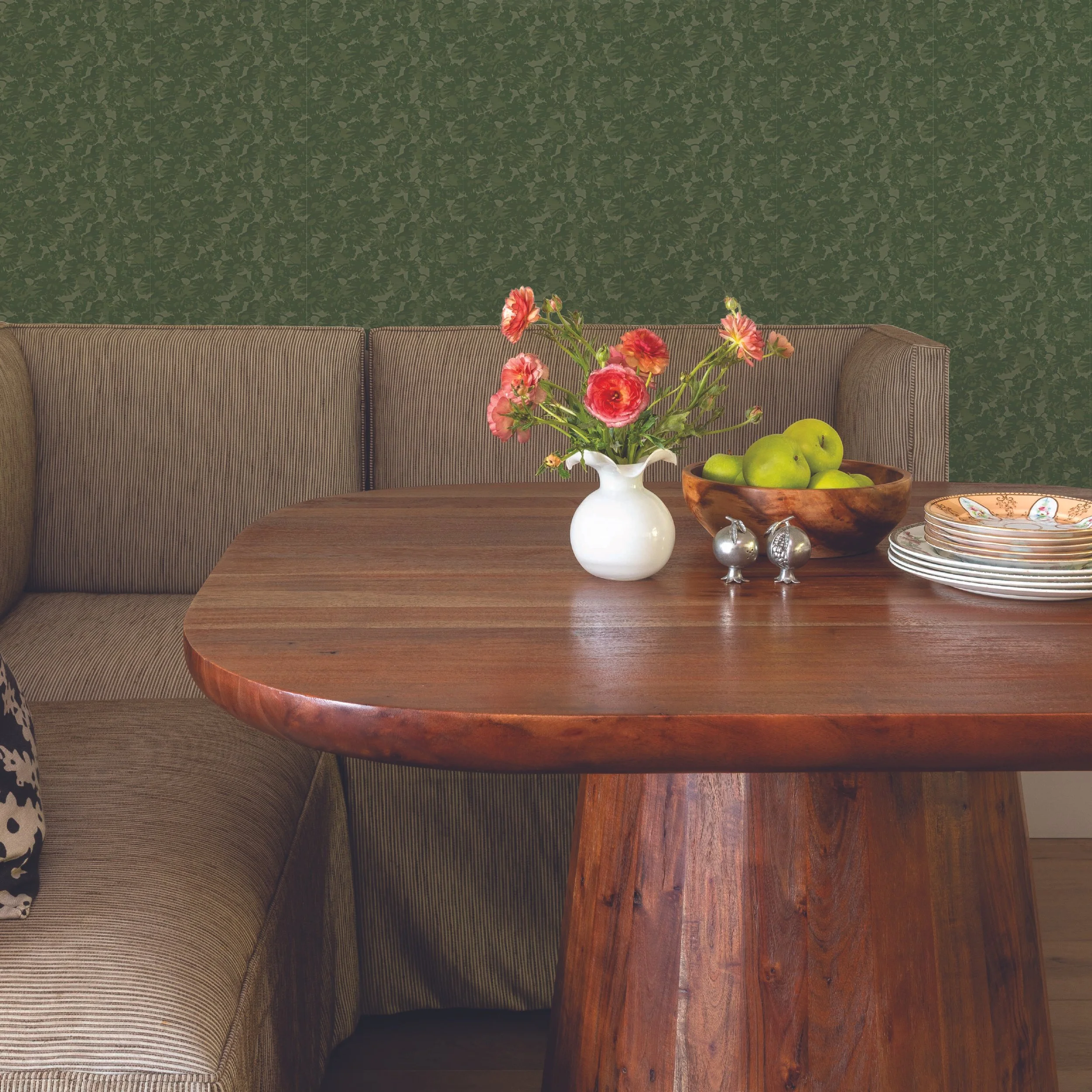 A dining setup with a wooden table, a white vase with pink flowers, and a wooden bowl with green apples. The background features a green patterned wallpaper and a brown sofa.