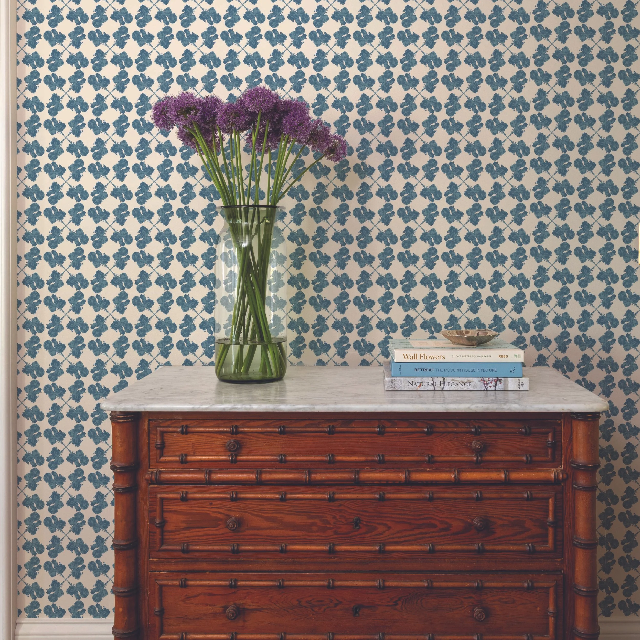 A bouquet of purple allium flowers in a green glass vase, placed on a white marble top table. Behind, there is a blue and white floral patterned wallpaper. To the right, on the table, there is a small stack of books, with titles about wall flowers, m