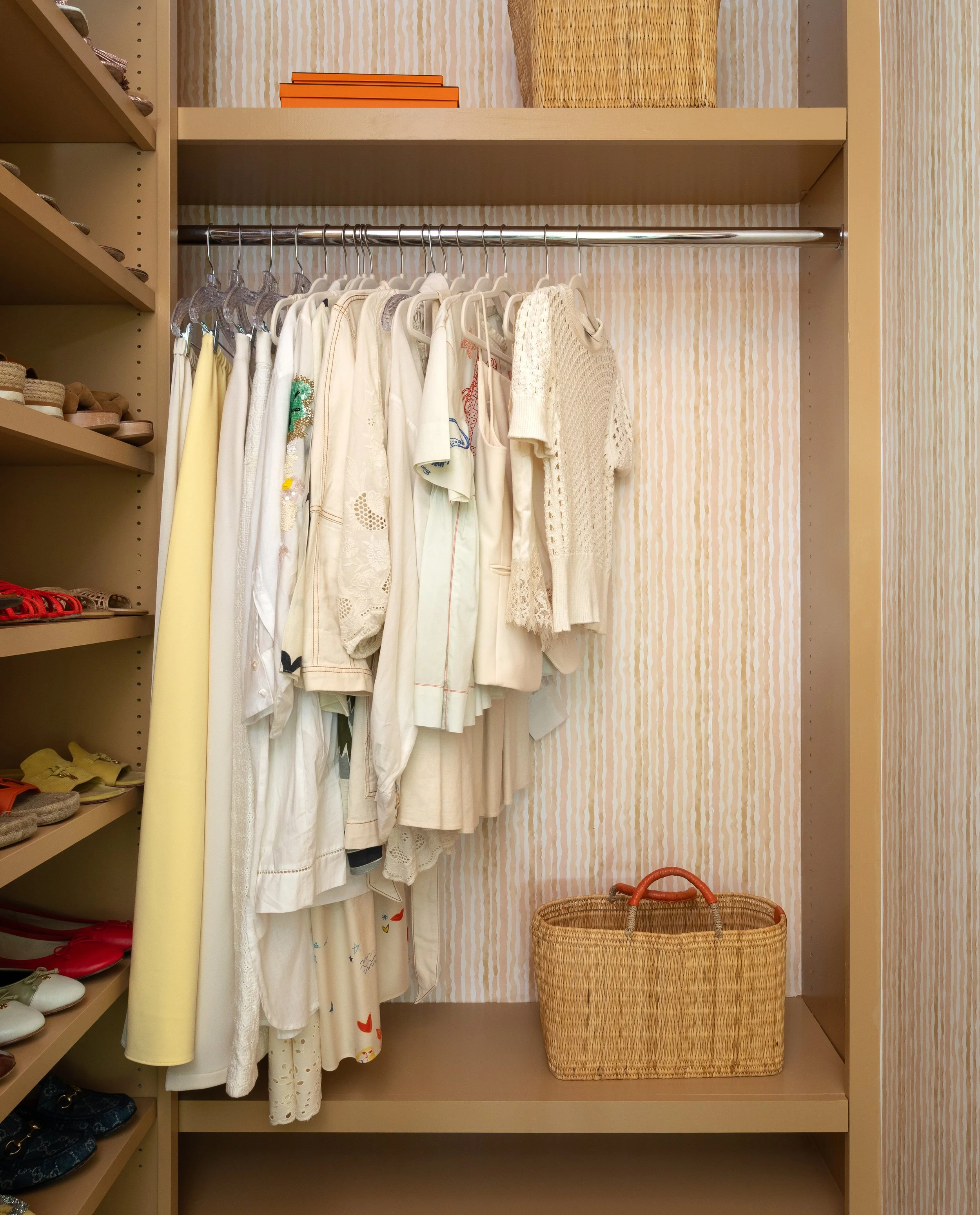 Closet with hanging vintage white and cream dresses, a woven basket on the floor, and shelves with shoes and orange boxes.